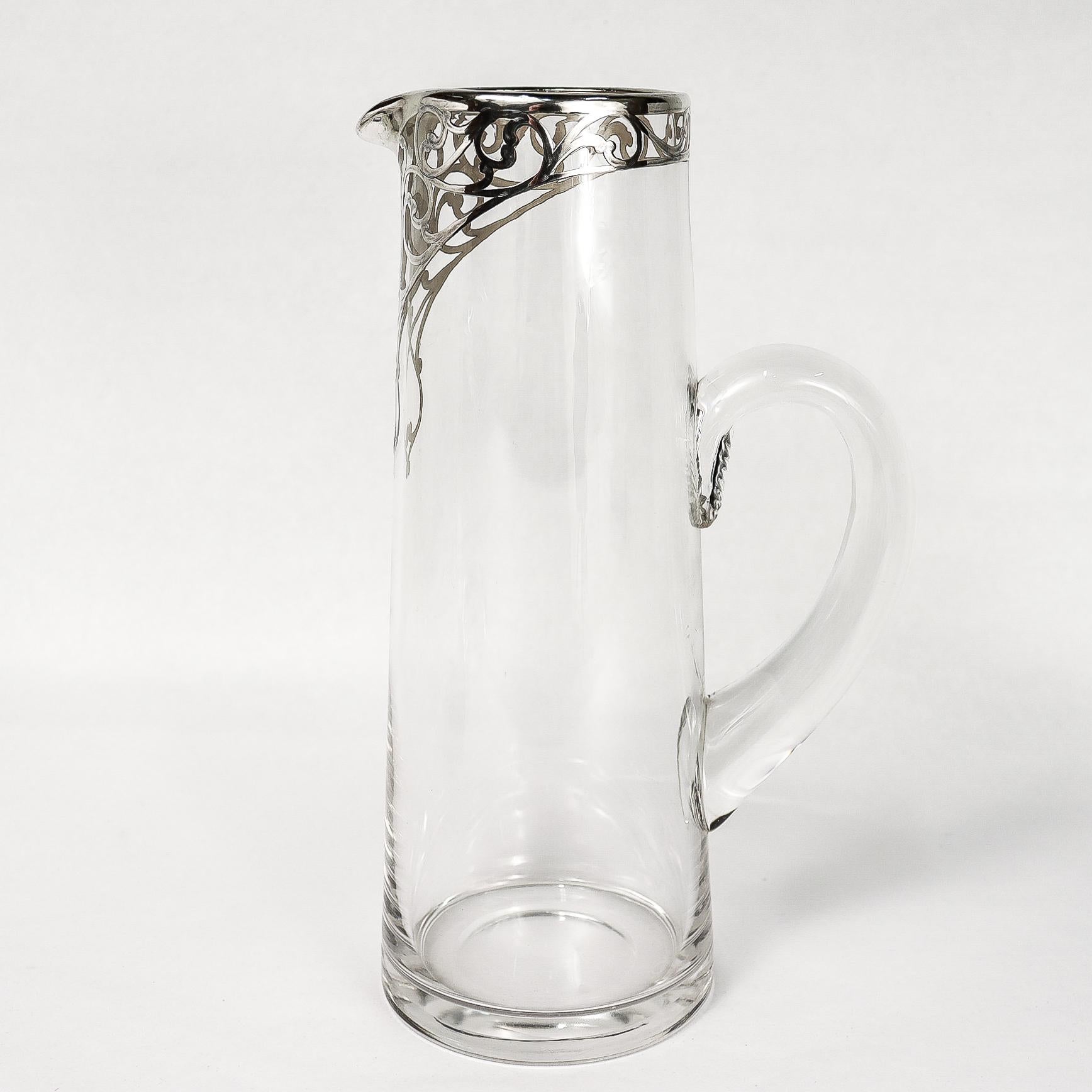 Una fina jarra antigua de cristal americano para cócteles o limonada.

De forma cilíndrica ahusada con asa aplicada.

Con incrustaciones de plata alrededor del borde y la parte superior de la jarra. La parte delantera de la jarra con una cartela