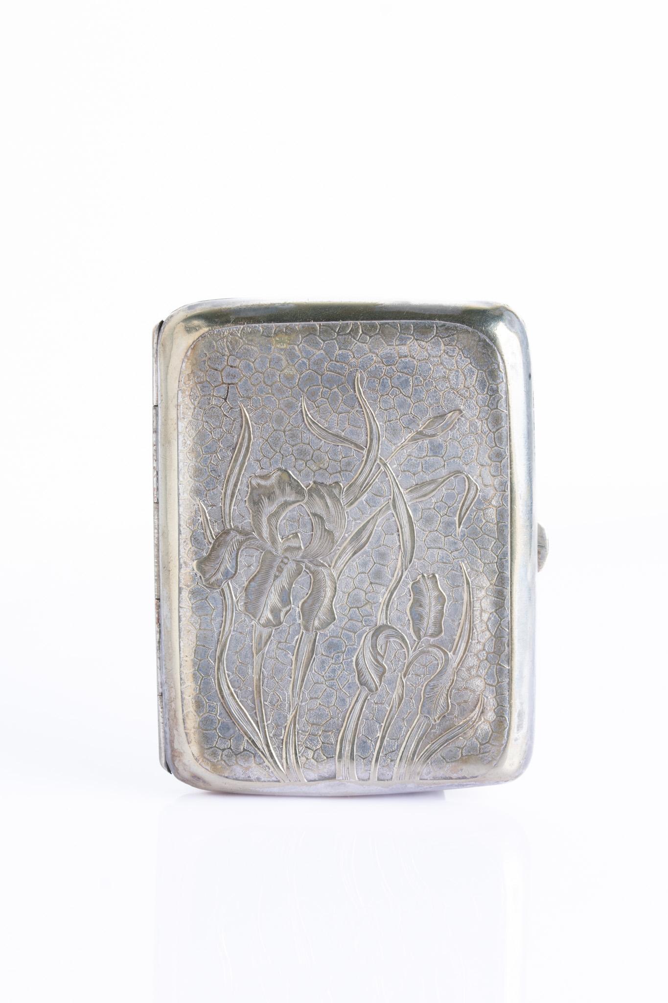 Un portasigarette decorativo in stile Art Nouveau placcato in argento, con fiori di iris in rilievo su uno sfondo martellato. L'iris - simbolo di fede, speranza, saggezza e rinnovamento - è stato uno dei motivi più amati dell'Art Nouveau e riflette