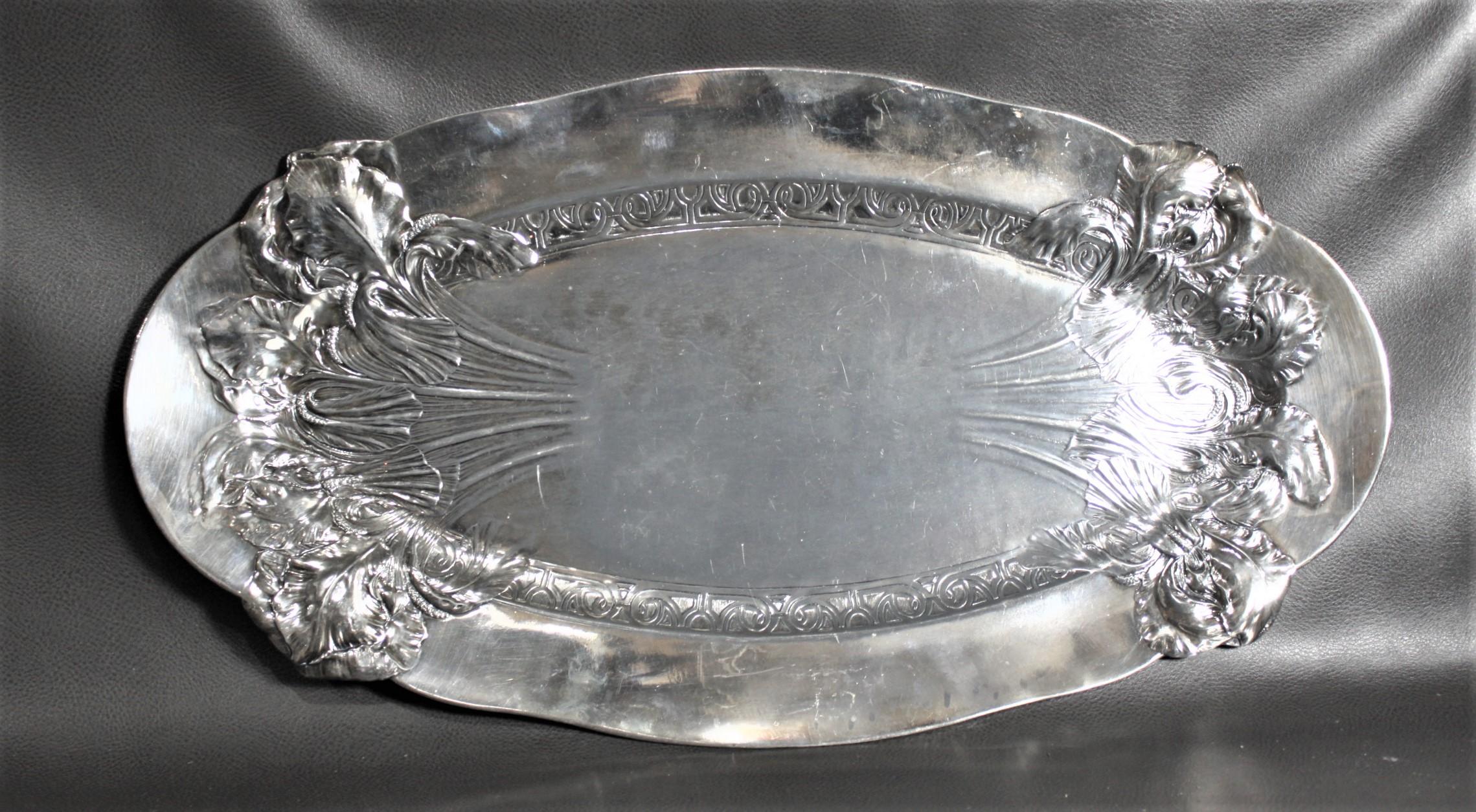 Esta antigua bandeja ovalada plateada para servir no lleva firma, pero se cree que fue fabricada en Inglaterra hacia 1920 en estilo Art Nouveau. La bandeja tiene forma oval alargada con borde alto y ancho y decoración floral estilizada en relieve