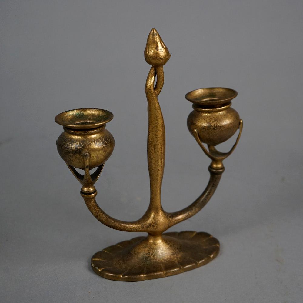 An antique Art Nouveau double candle stick by Tiffany Studios features Dore bronze construction with central stylized fleur de lis or flame torchiere column having two arms, each terminating in urn form candle sockets raised on three prongs, maker