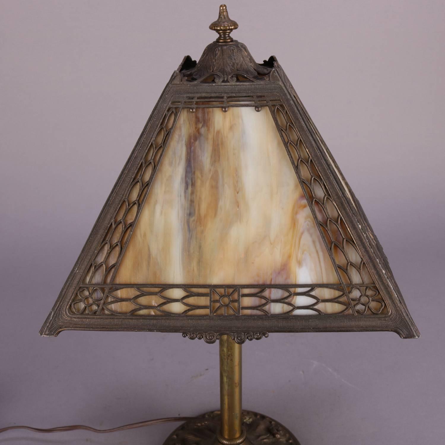 Antique Arts & Crafts Miller petite table lamp features cast base with high relief foliate and floral design, four panel shade with pierced filigree shade frame with foliate border and central stylized flower and housing four slag glass panels,