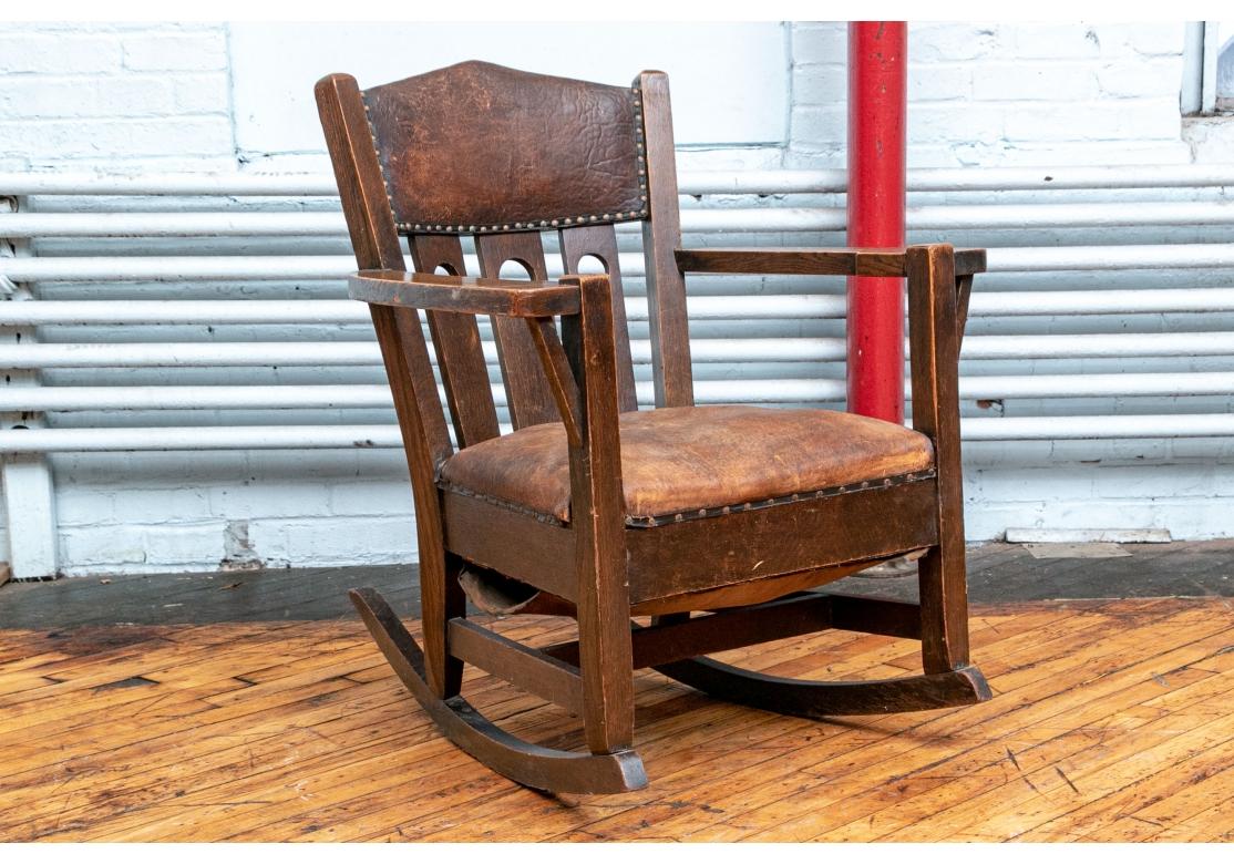 A tightly made and comfortable Period Arts and Crafts Period Rocking Chair. With a triangular leather upholstered deep crest rail with nailhead trim. The triple slat back with carved open circles. With classic shaped flat and wide arms with straight