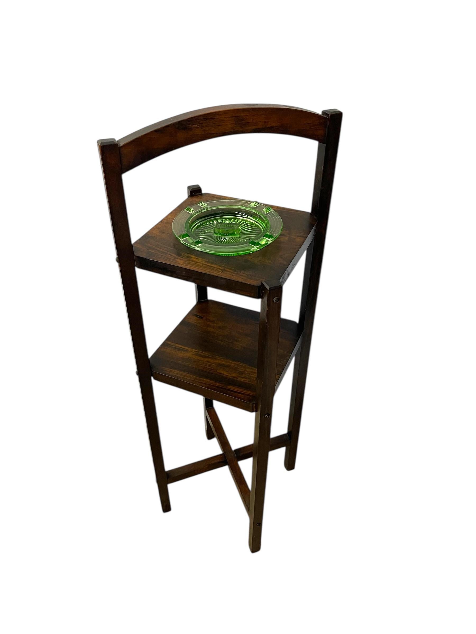 This antique two-tiered smokers table reflects the simple elegance of the Arts and Crafts movement. Constructed from solid wood with a warm, dark-stained finish, the piece features a tall, narrow frame with an arched backrest, square legs, and