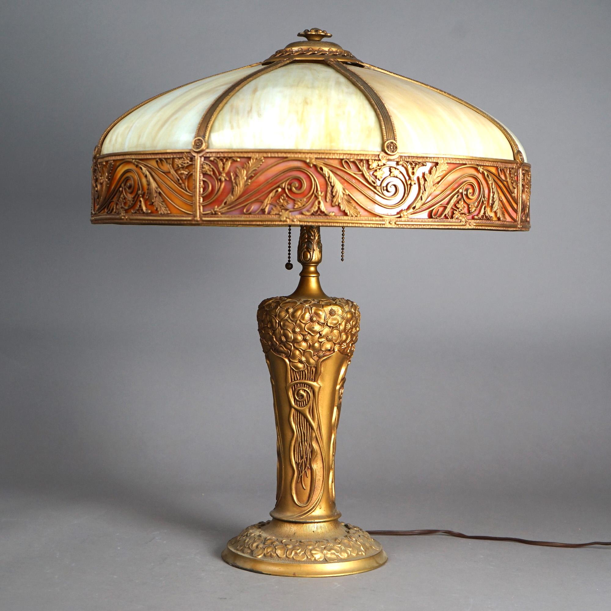 An antique Arts and Crafts into Art Nouveau table lamp by Royal offers cast shade with scroll and foliate elements housing two tone slag glass over cast double socket base with foliate elements, c1920

Measures - 24.5
