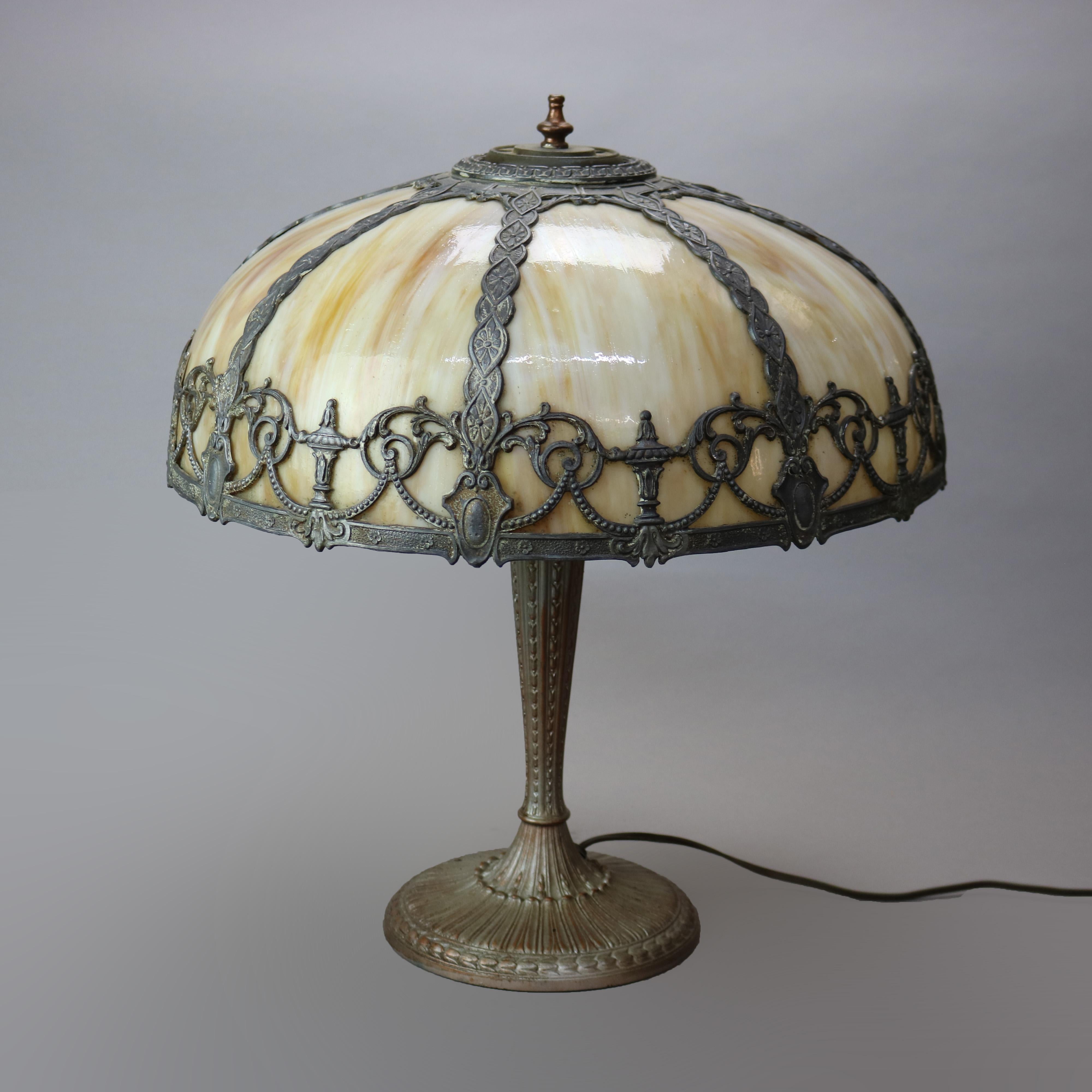 Antigua lámpara de sobremesa Arts & Crafts a la manera de Bradley & Hubbard, con pantalla en forma de cúpula y pantalla fundida con elementos de urna, guirnalda, escudo y volutas foliadas, que alberga paneles de vidrio de escoria curvado sobre base