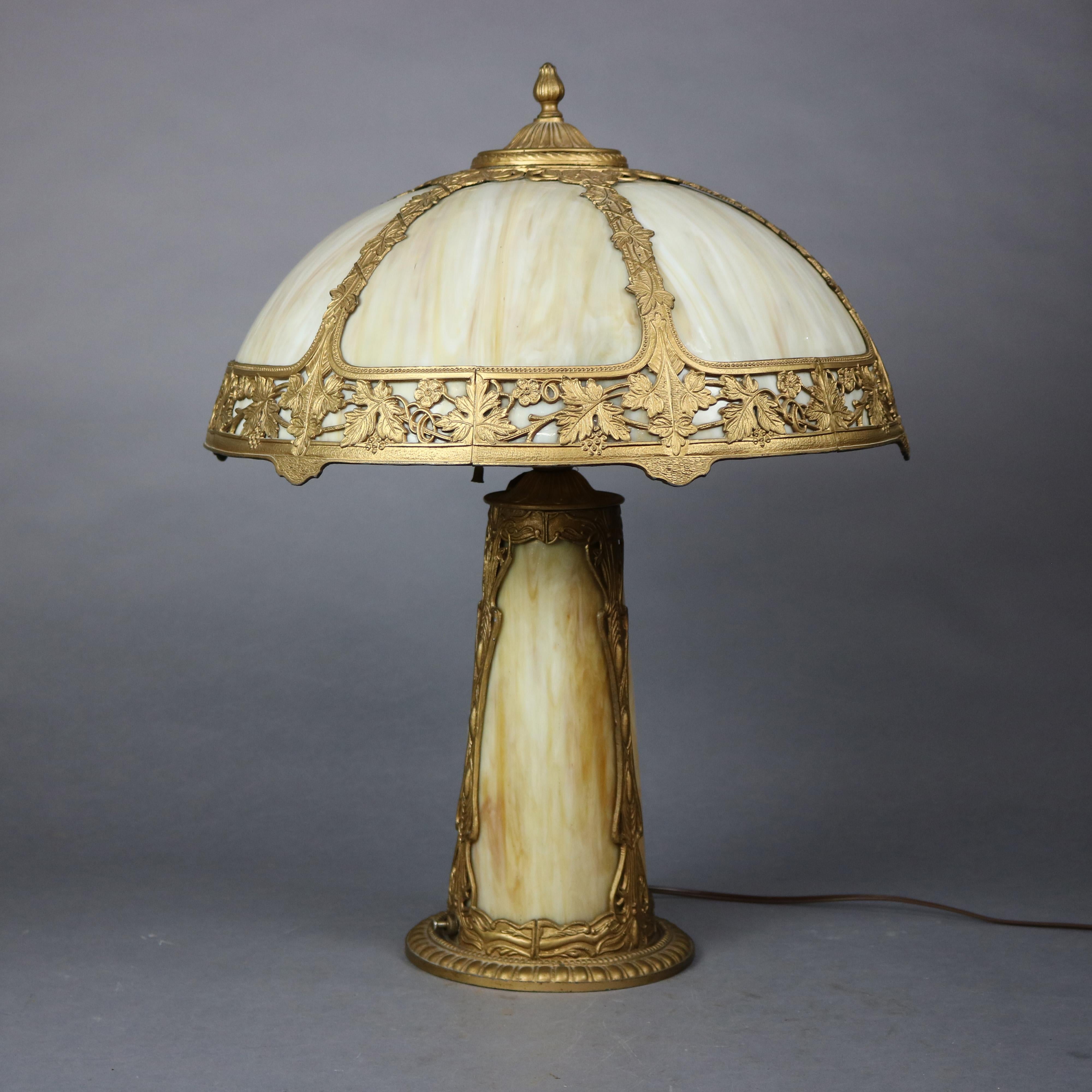 An antique Arts and Crafts table lamp in the manner of Bradley and Hubbard offers dome shade having cast frame with leaf and berry vine motif housing curved slag glass panels surmounting double socket flared base, circa 1910

Measures - 23.5''H x