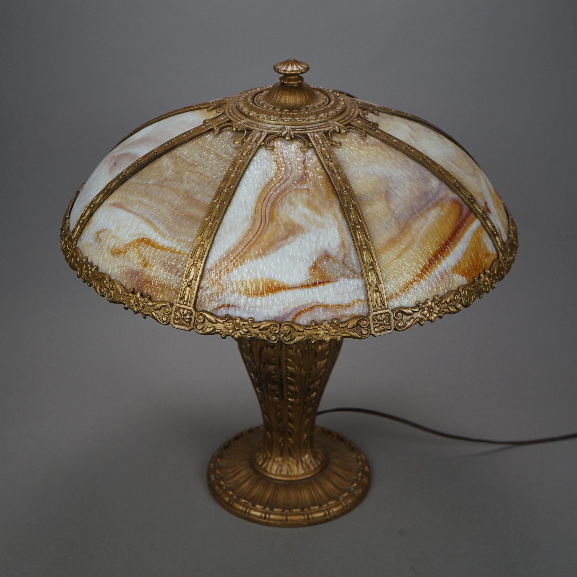 An antique Arts & Crafts table lamp in the manner of Bradley and Hubbard offers cast frame with floral and foliate decoration and housing bent caramel slag glass panels over triple socket cast urn form base with foliate elements, c1920

Measures-