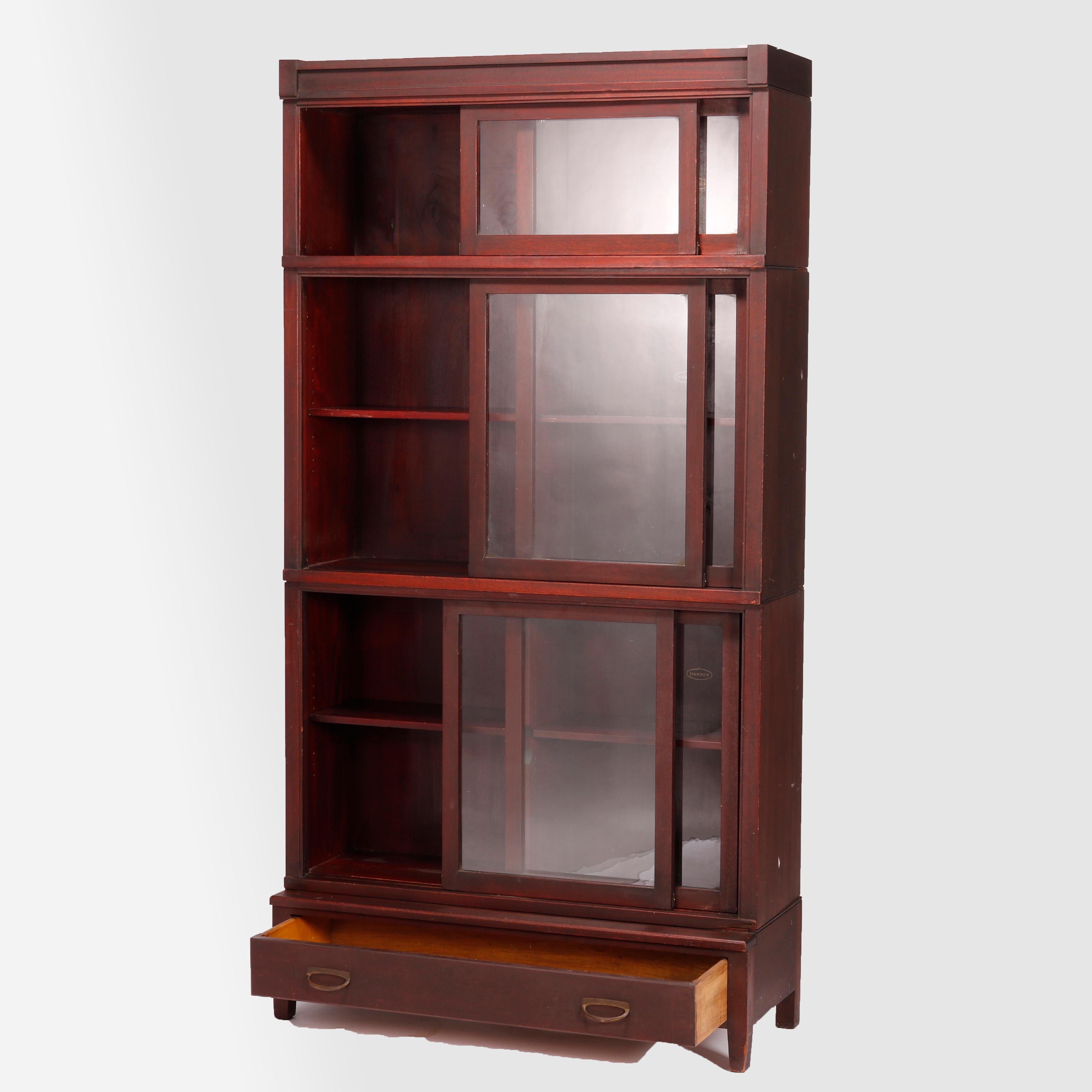 An antique Arts and Crafts bookcase by Danner and retailed by Paine of Boston offers mahogany construction with three stacks each having sliding glass doors 
 and raised on single drawer base with straight and square legs, maker and retailer labels