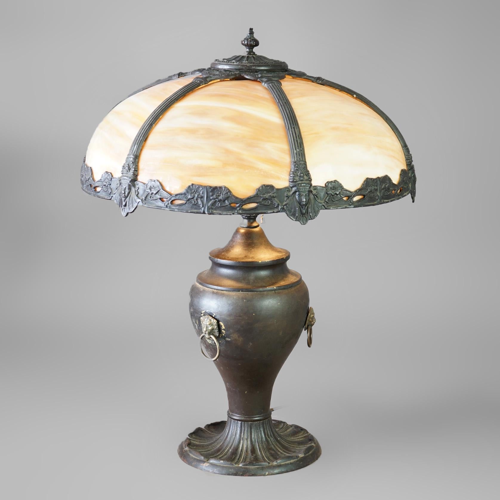 An antique Arts and Crafts table lamp in the manner of Bradley and Hubbard offers dome form shade with cast metal frame having foliate and Egyptian mask elements and housing curved slag glass panels; over cast urn form base with figural handles,