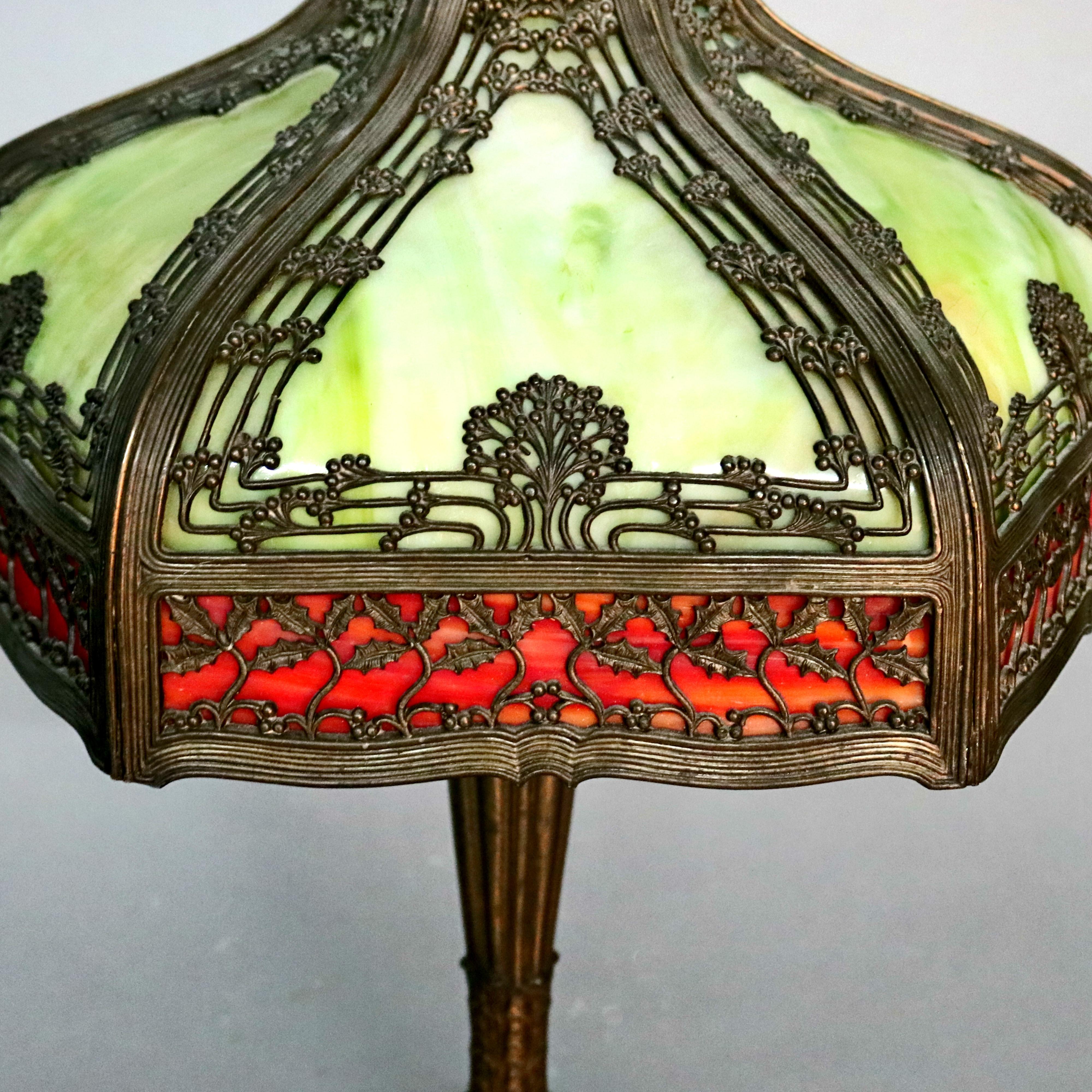 An antique Arts & Crafts table lamp by Royal Co. offers cast bronze foliate and Tree of Life filigree shade housing six moss green slag glass and bordered with six red-orange slag glass panels surmounting foliate cast metal base having two