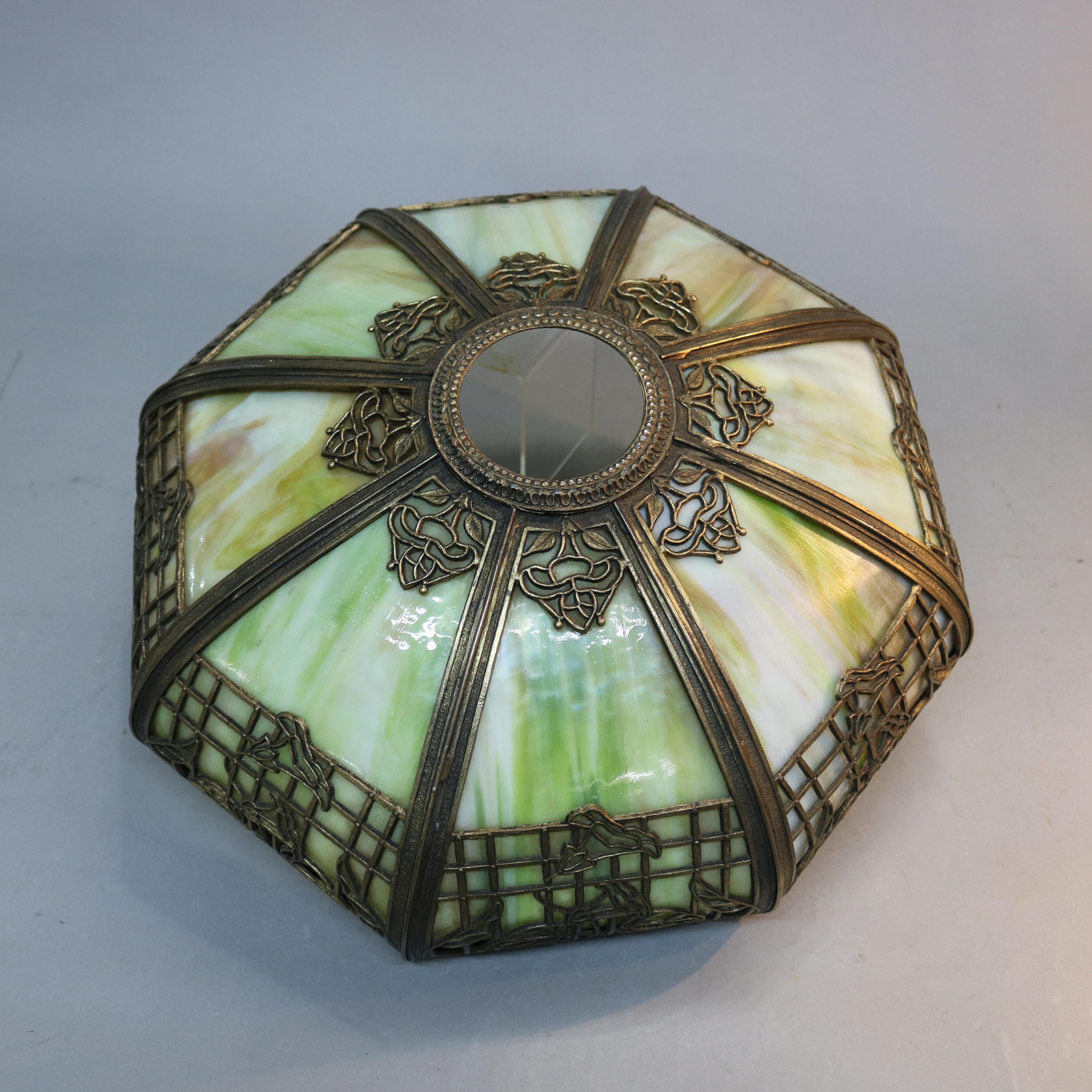 An antique Arts & Crafts table lam offers paneled cast shade with floral and foliate filigree designing housing bent slag glass surmounting cast base, circa 1920.

Measures: 22