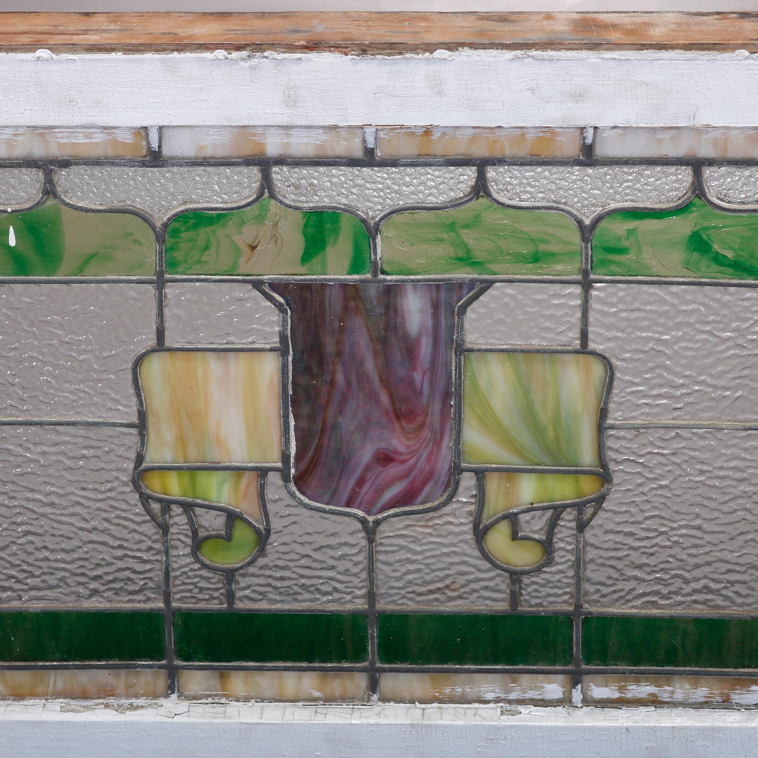 An antique Arts & Crafts leaded glass window in the manner of Frank Lloyd Wright offers slag and colorless glass panel having central shield and flanking stylized floral elements, seated in original wood frame, circa 1910

Measures: 23