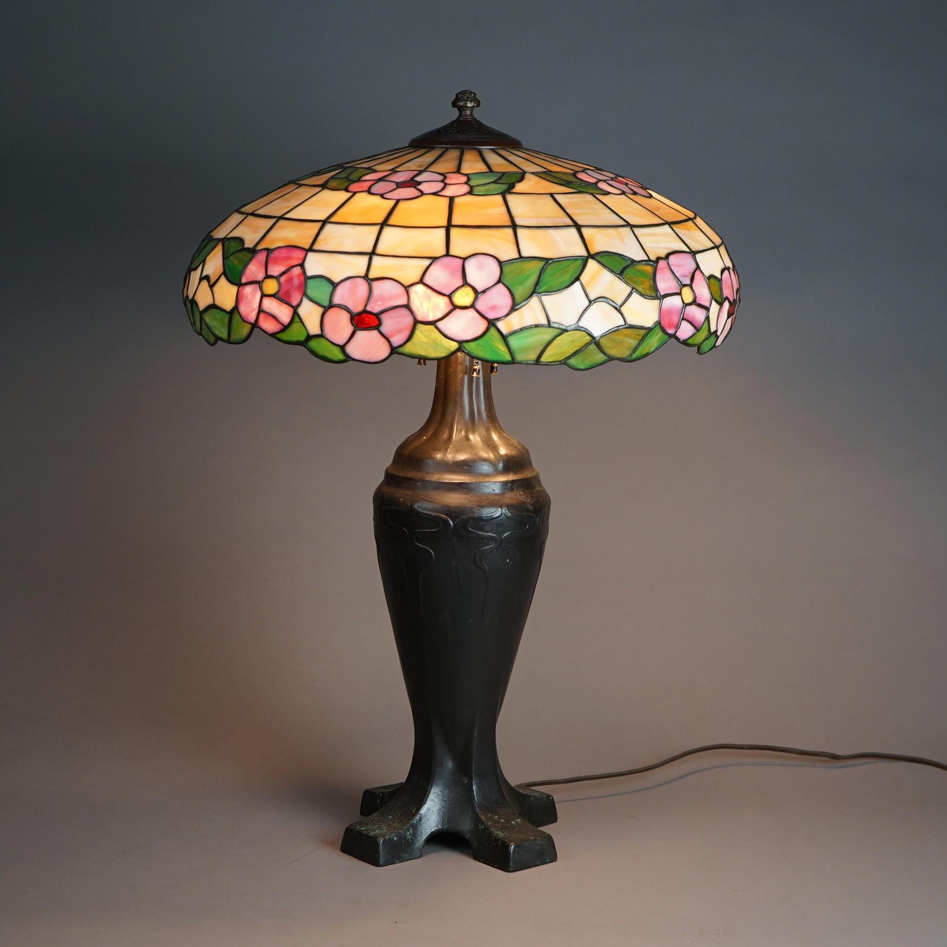 An antique Arts and Crafts table lamp in the manner of Handel offers leaded stained and slag glass shade with flowers over urn form cast bronze footed base with stylized embossed foliate decoration and four feet, c1920

Measures- 26.5''H x 19''W x