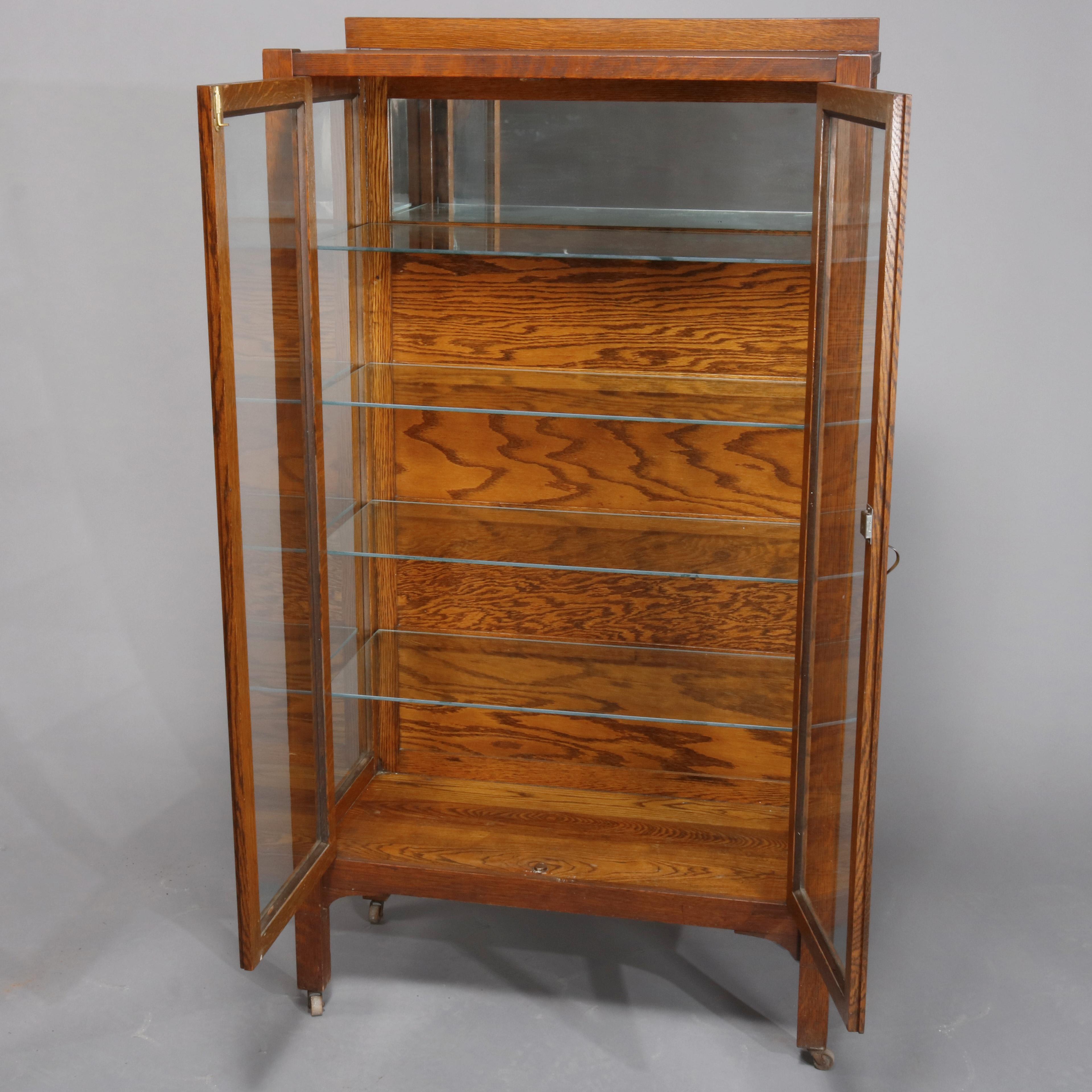 An antique Arts & Crafts mission oak china cabinet offers oak construction with low backsplash surmounting case having glass sides and double glass doors opening to partially mirrored and shelved interior, raised on straight and square legs with