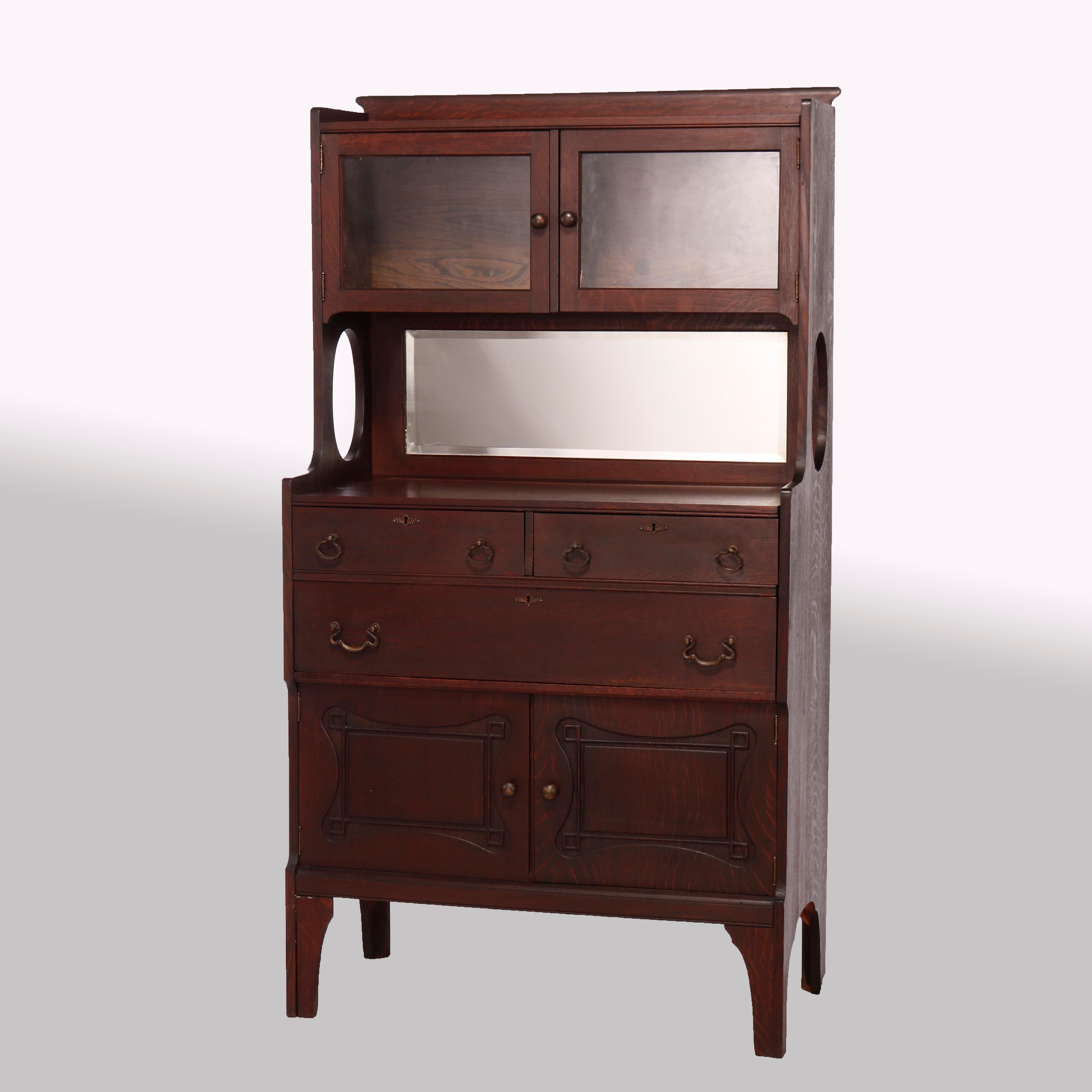 An antique Arts and Crafts Mission curio server offers quarter sawn oak construction with upper double glass door cabinet surmounting mirrored backsplash over lower case with double drawers over single long drawer and two blind cabinets having doors