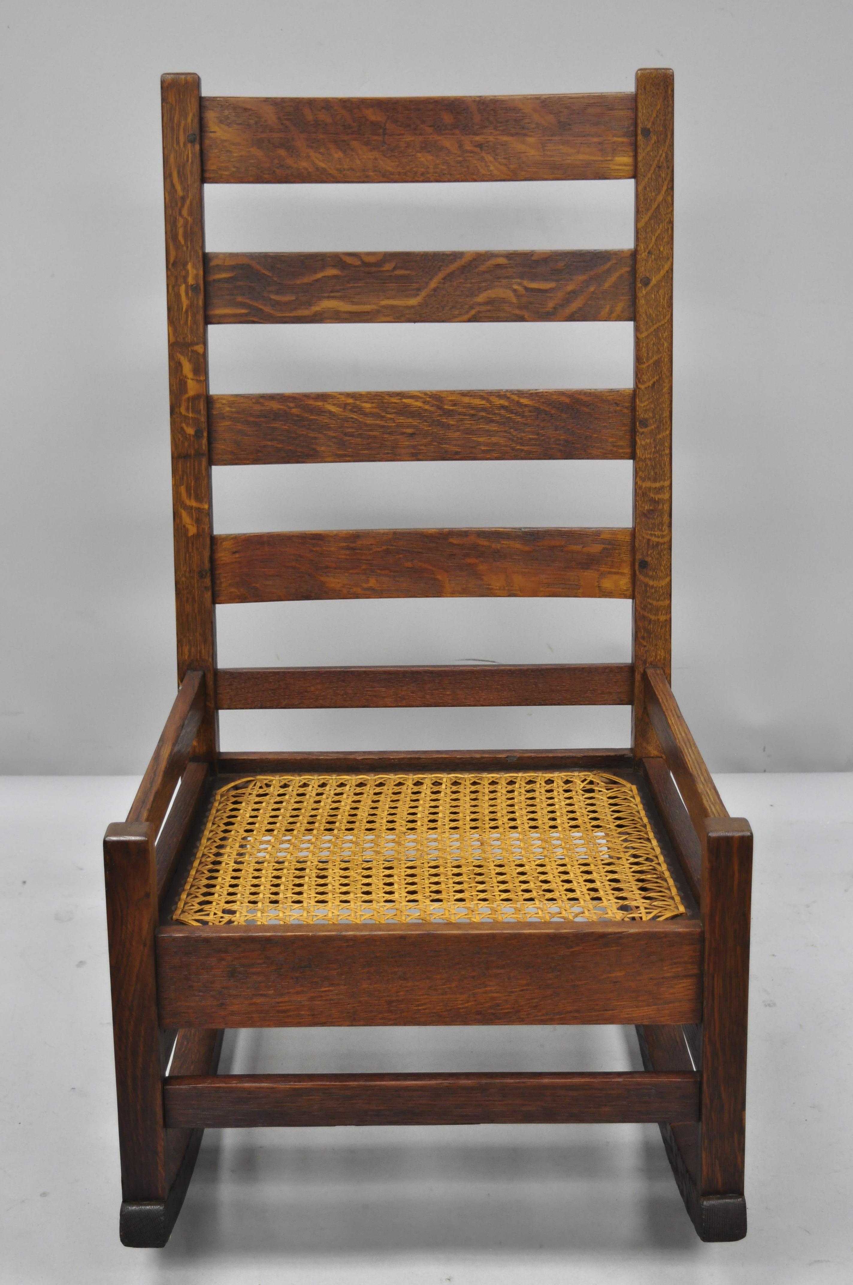 Antique Arts & Crafts mission oak ladder back hip rail rocking chair. Cane seat, ladder back exposed dowel joinery, solid wood construction, beautiful wood grain, very nice antique item, unsigned but possibly by Stickley, circa 1900. Measurements: