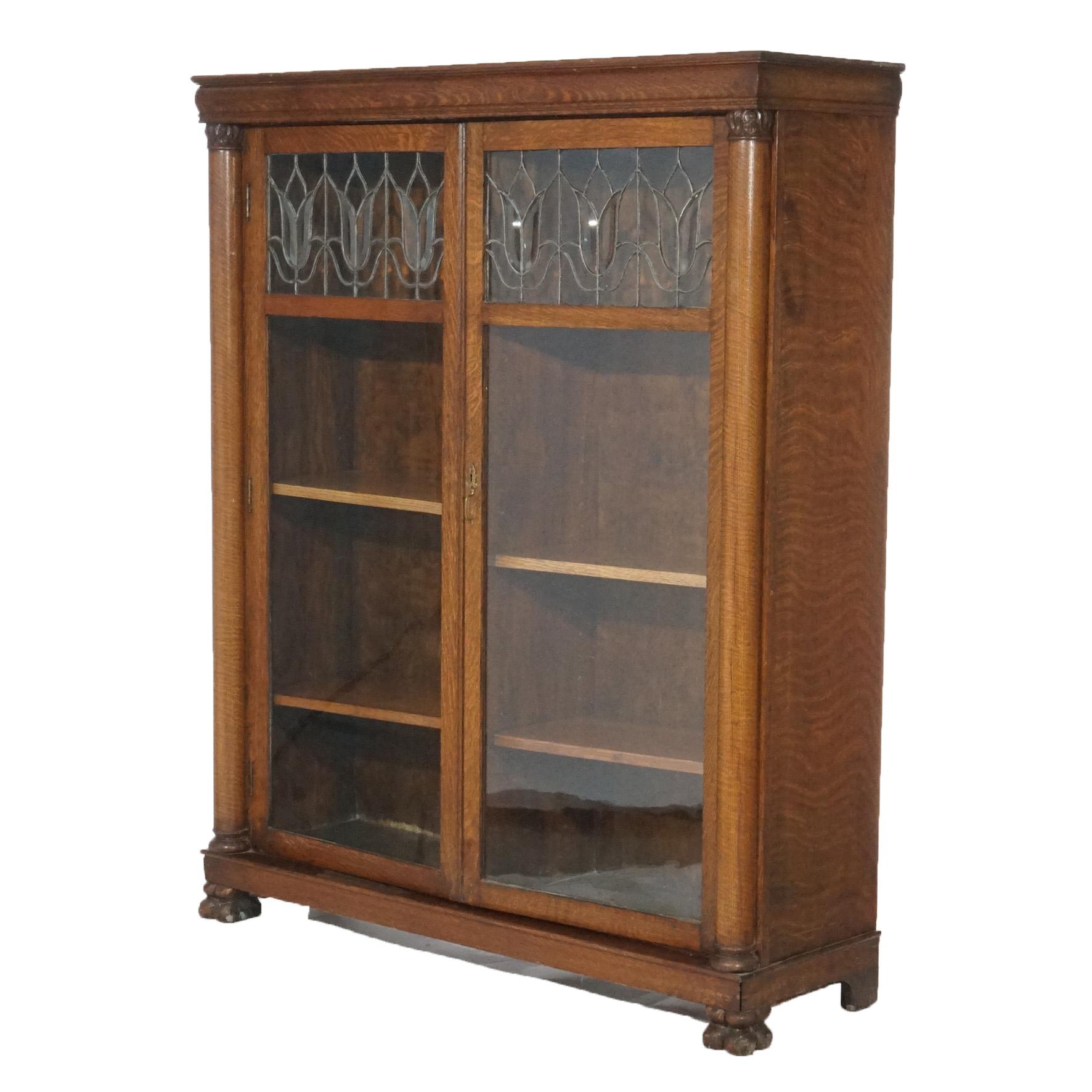 An antique Arts and Crafts Mission bookcase offers quarter sawn oak construction with double glass doors having leaded glass upper panels opening to divided adjustable shelf interior, flanking full columns and raised on stylized carved paw feet,