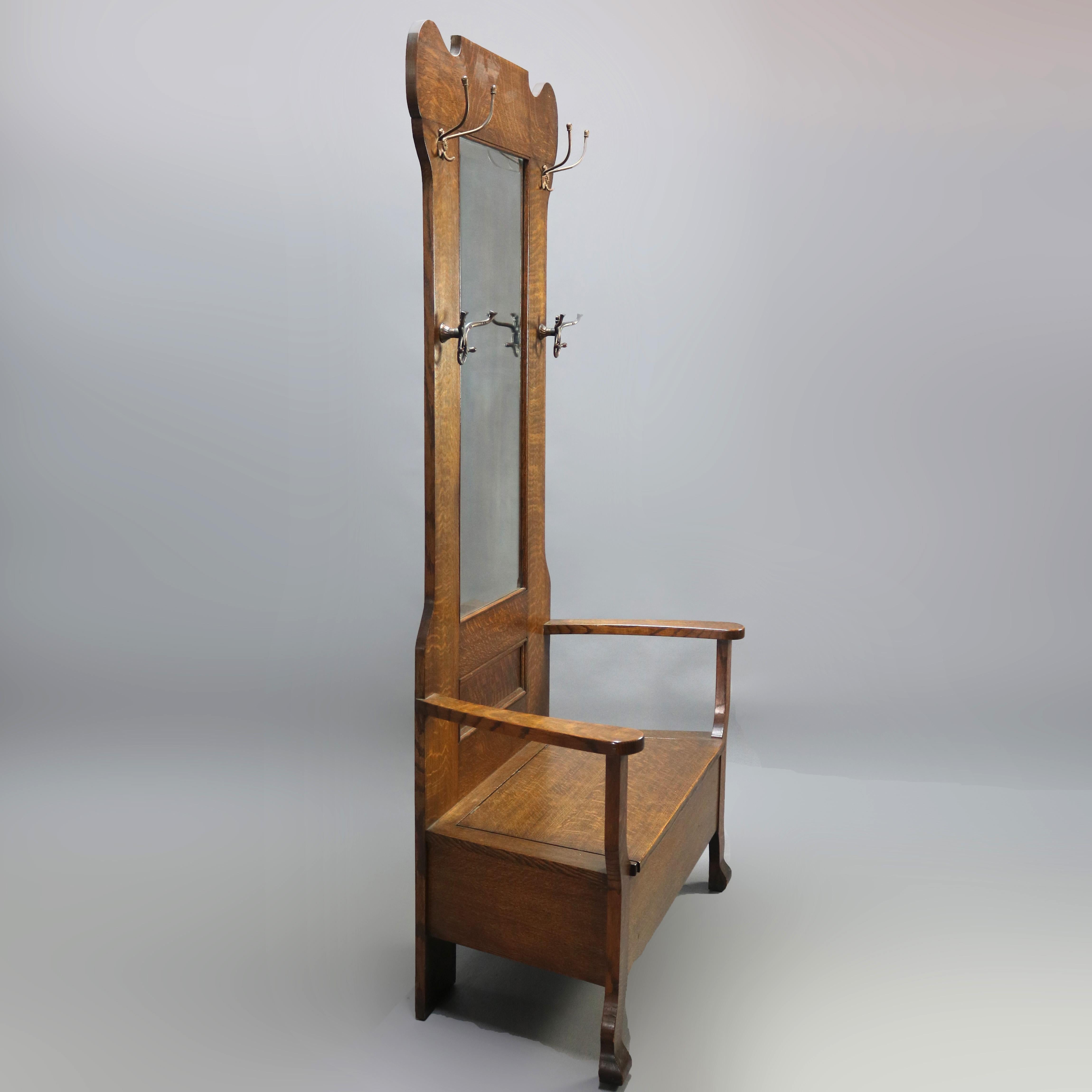 An antique Arts & Crafts Mission oak hall seat offers shaped frame with central mirror and flanking coat/hat hooks surmounting lift top seat with level arms, circa 1900.

Measures - 79.5
