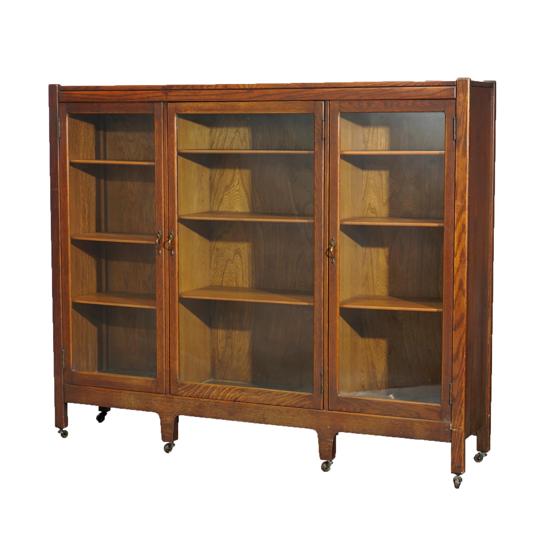 An antique Arts and Crafts enclosed bookcase offers quarter sawn oak construction with three glass doors opening to divided and adjustable shelved interiors, raised on square and straight legs with casters, c1910

Measures - 49.75'' H x 59.25'' W