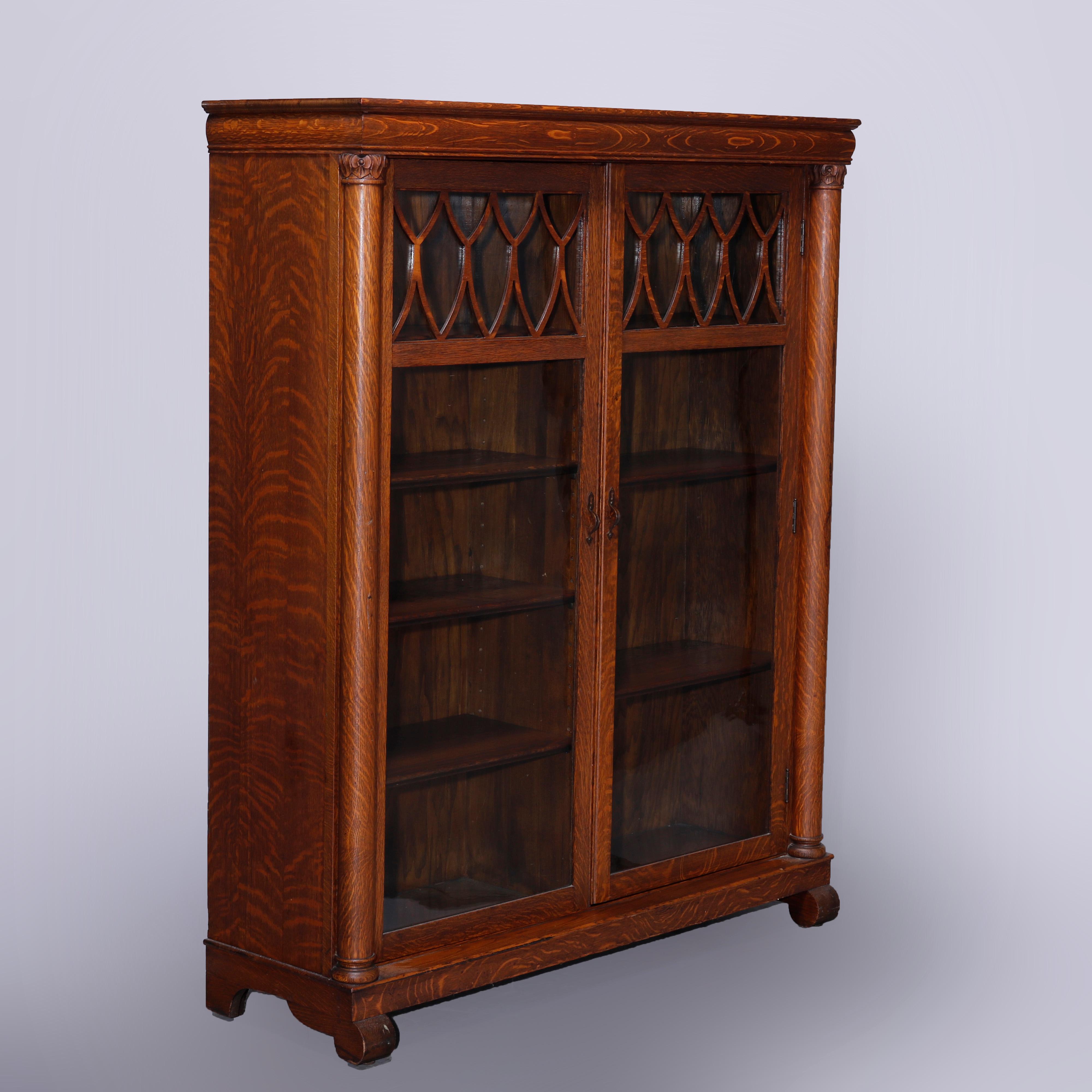 An antique Arts & Crafts enclosed bookcase offers quarter sawn oak construction with double glass doors having cut-out stylized shield upper opening to divided and adjustable shelf interior, flanked by full Corinthian column supports having carved