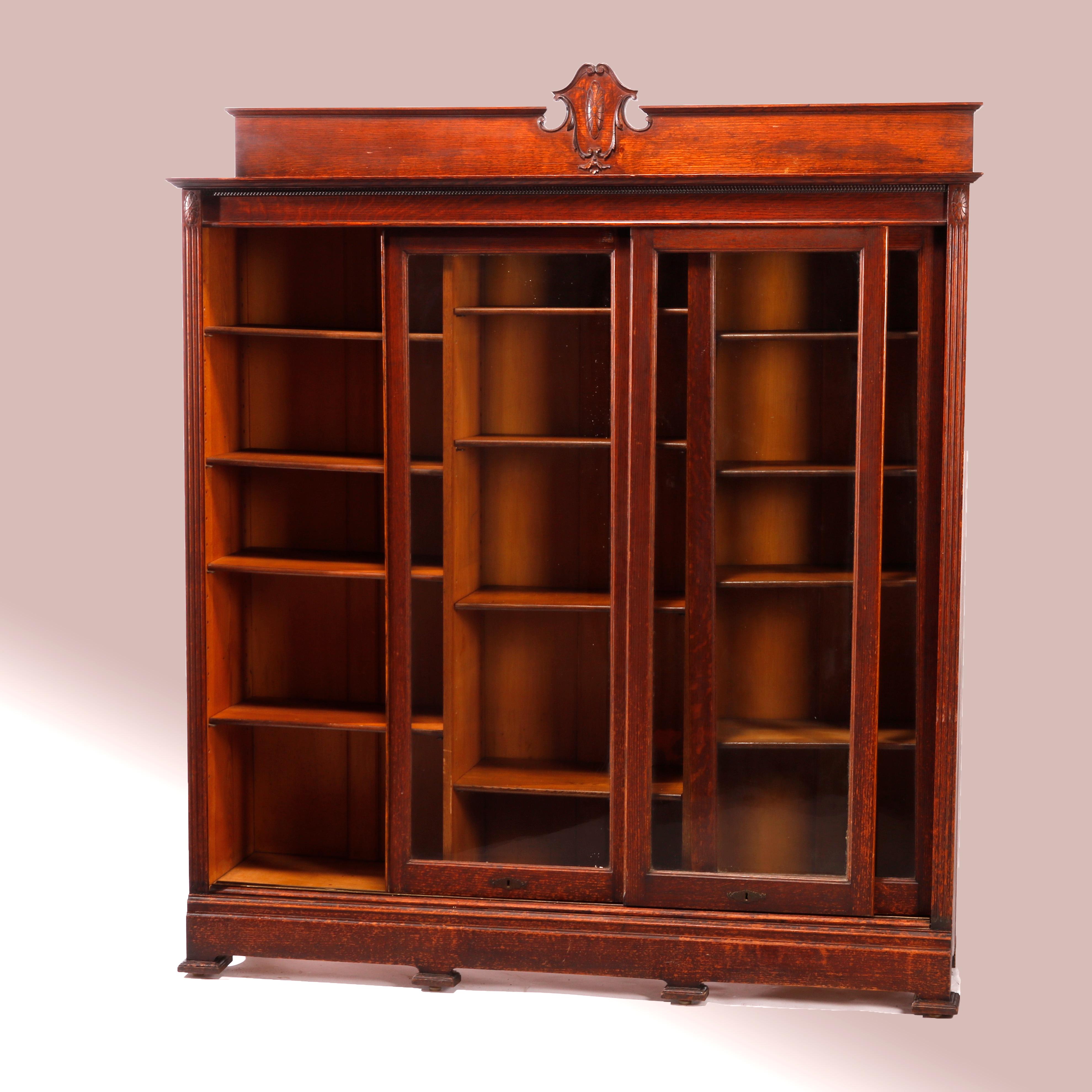 An antique Arts & Crafts bookcase offers quarter sawn oak construction with upper having backsplash with carved shield form crest over triple sliding glass door opening to adjustable shelf divided interior, raised on square feet, c1910

Measures -