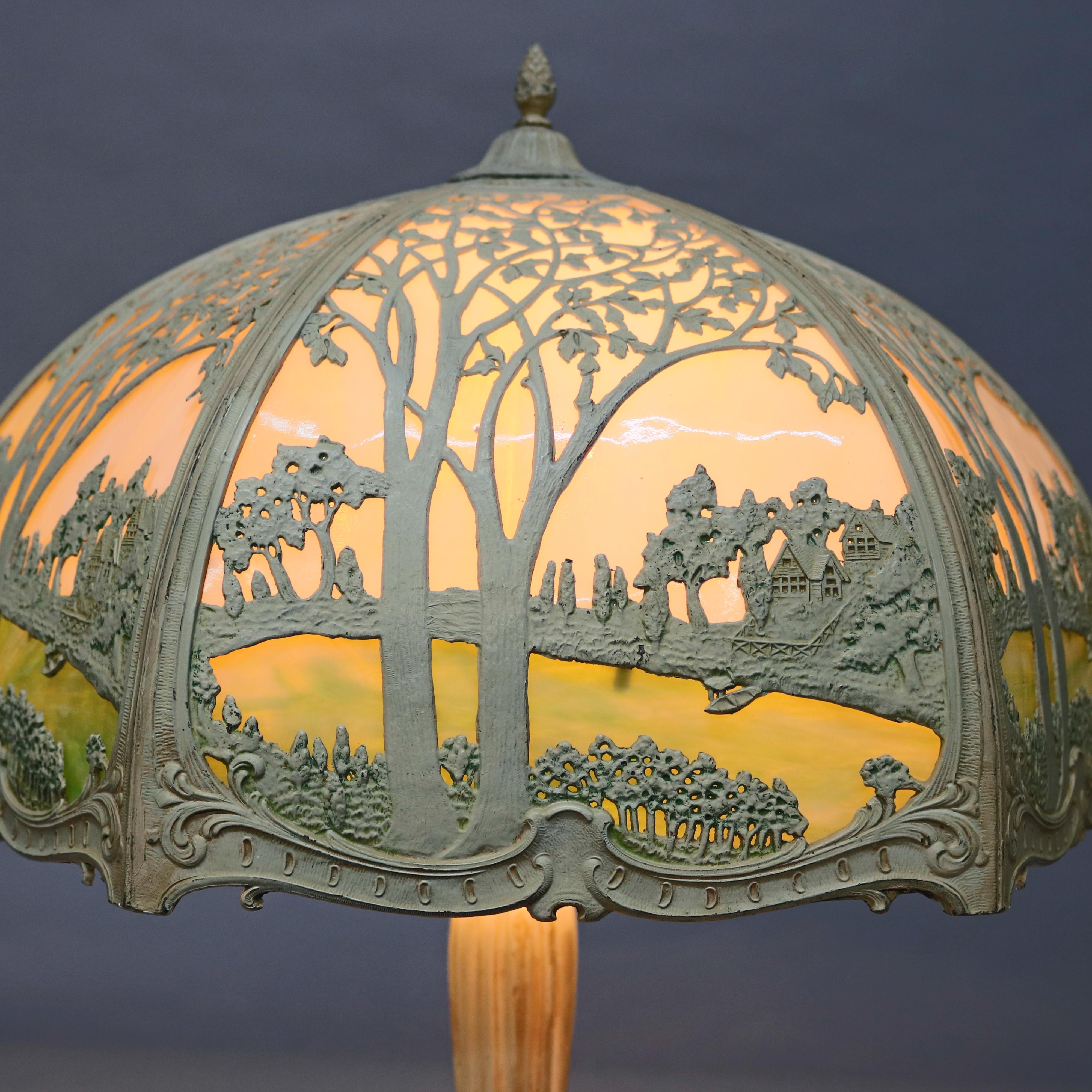 An antique Arts & Crafts oversized lamp by Miller offers cast metal filigree shade with landscape scene housing two-toned bent slag glass surmounting triple socket scrolled acanthus foliate base, circa 1920

Measures: 25