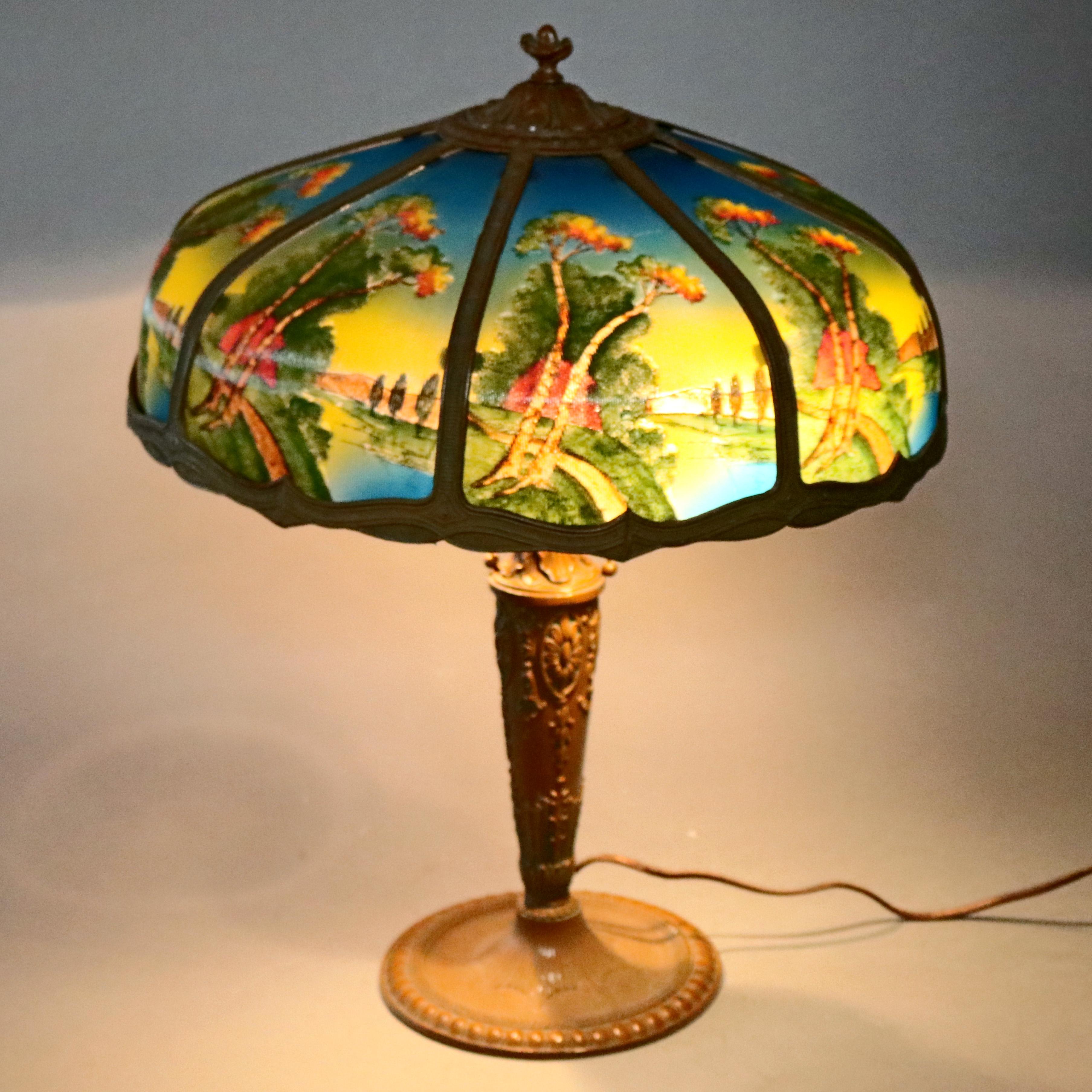 An antique Arts & Crafts Pittsburgh School table lamp offers a cast shade housing reverse painted ribbed glass panels depicting country side river scene and surmounting cast urn form base having embossed floral elements with inverted bellflowers and