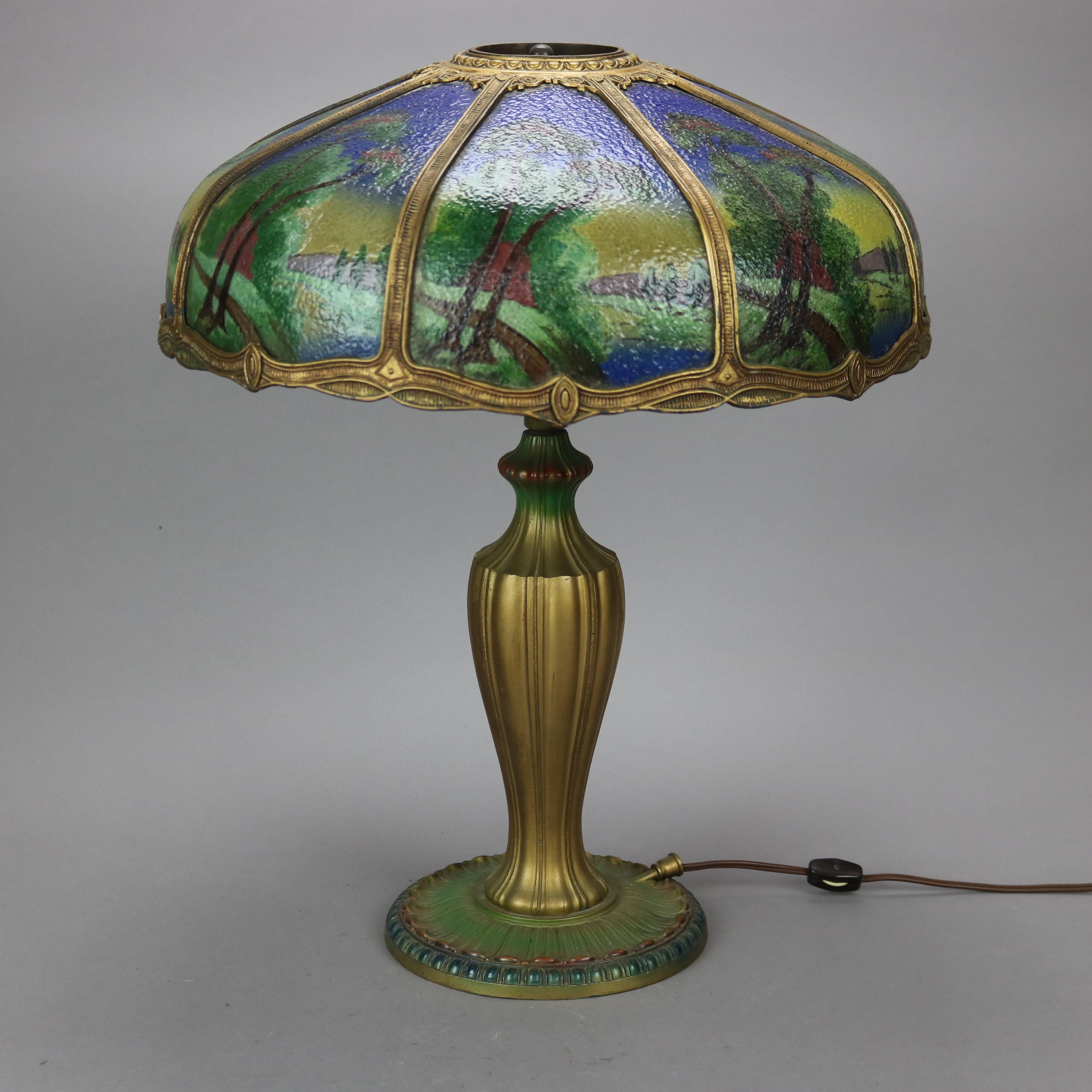 An antique Arts and Crafts table lamp in the manner of Bradley and Hubbard offers cast frame housing bent glass and hand painted glass panels having countryside scene over double socket cast urn form base, c1920

Measures - 21'' H x 17.5'' W x