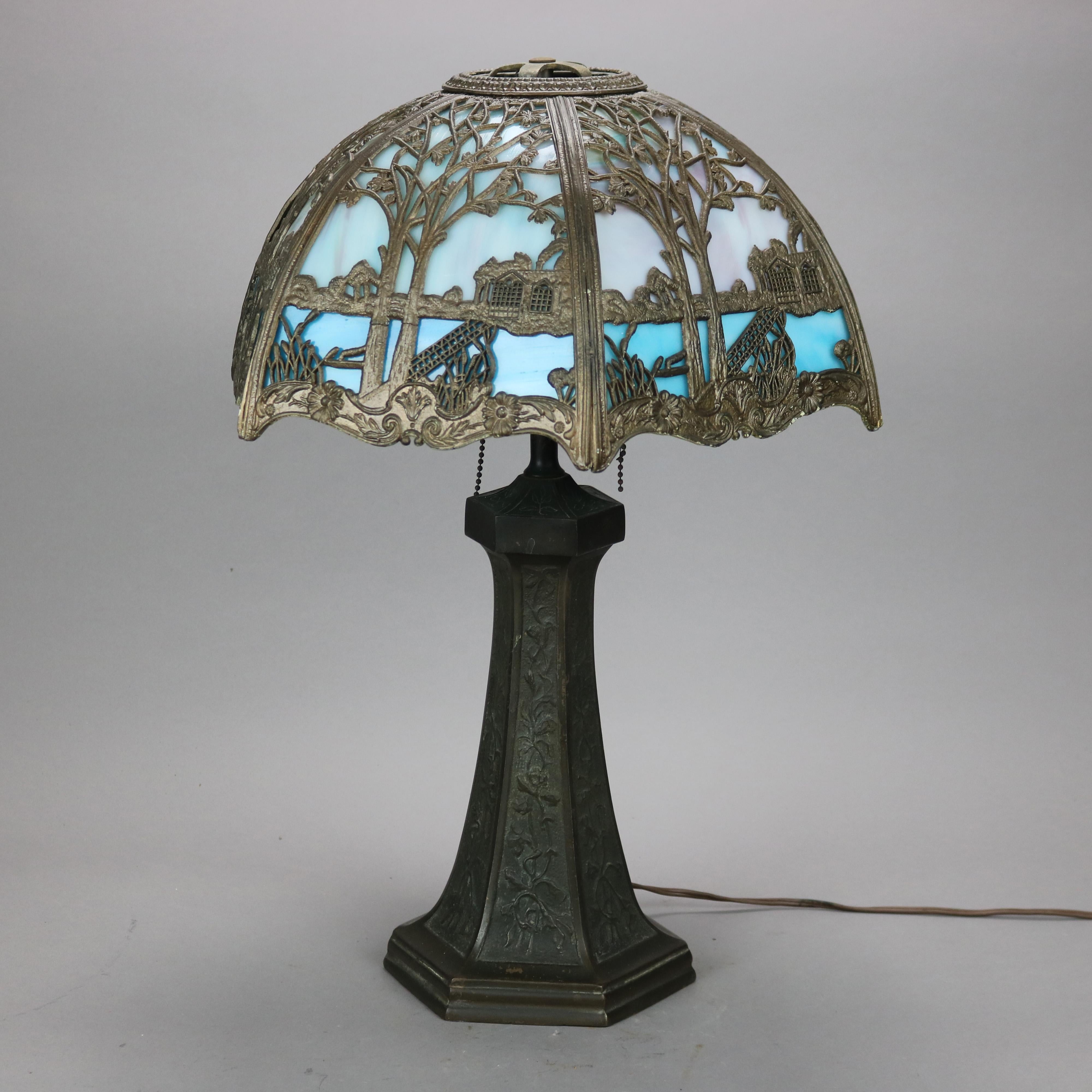 An antique Arts & Crafts table lamp in the manner of Bradley and Hubbard offers scenic filigree shade with landscape scene having structure and bridge, housing two tone bent slag glass panels over double socket faceted and flared base with foliate