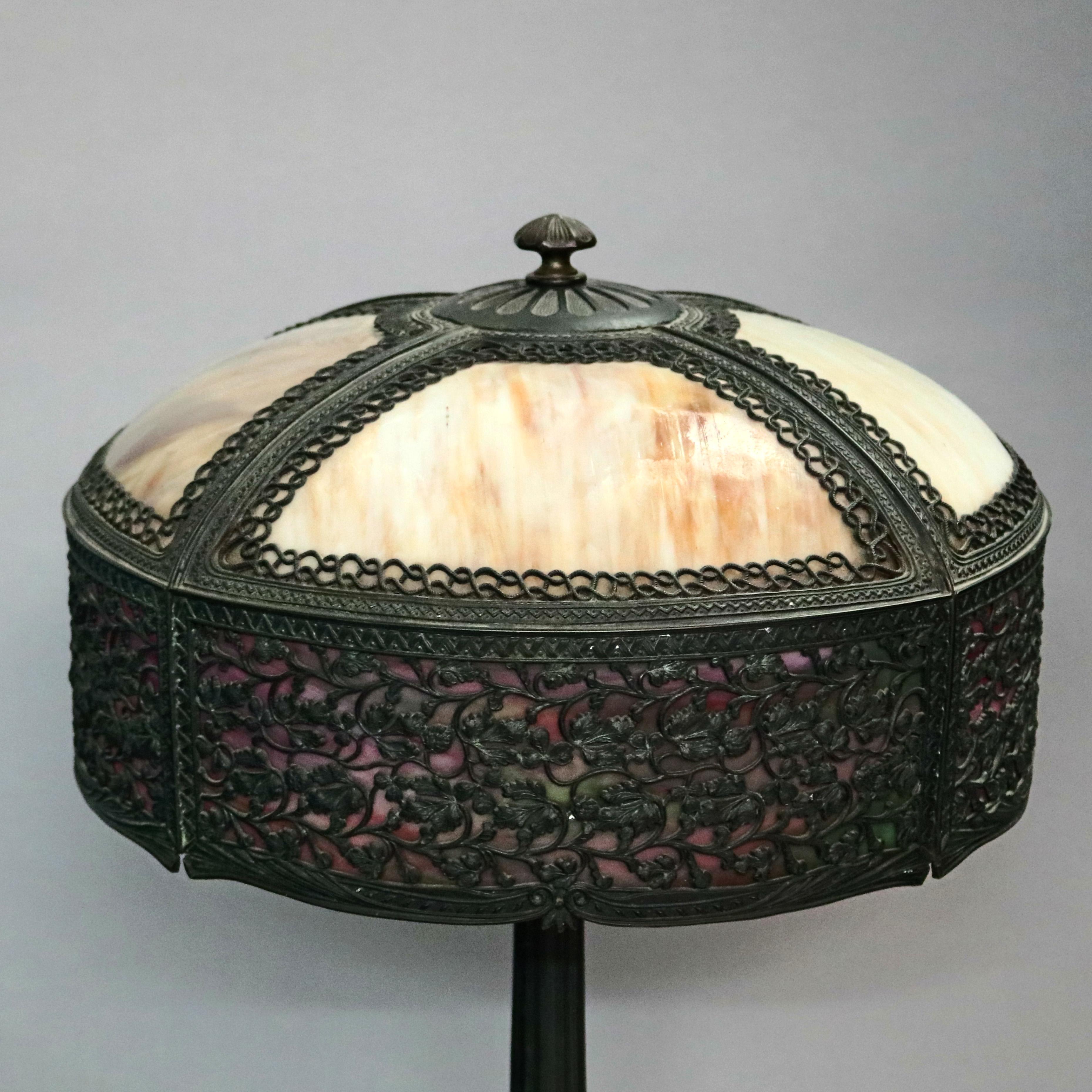 An antique Arts & Crafts table lamp in the Bradley & Hubbard style offers shade with cast frame having pierced foliate rim housing two-toned slag glass and surmounting cast foliate form base having two independently controlled sockets, circa