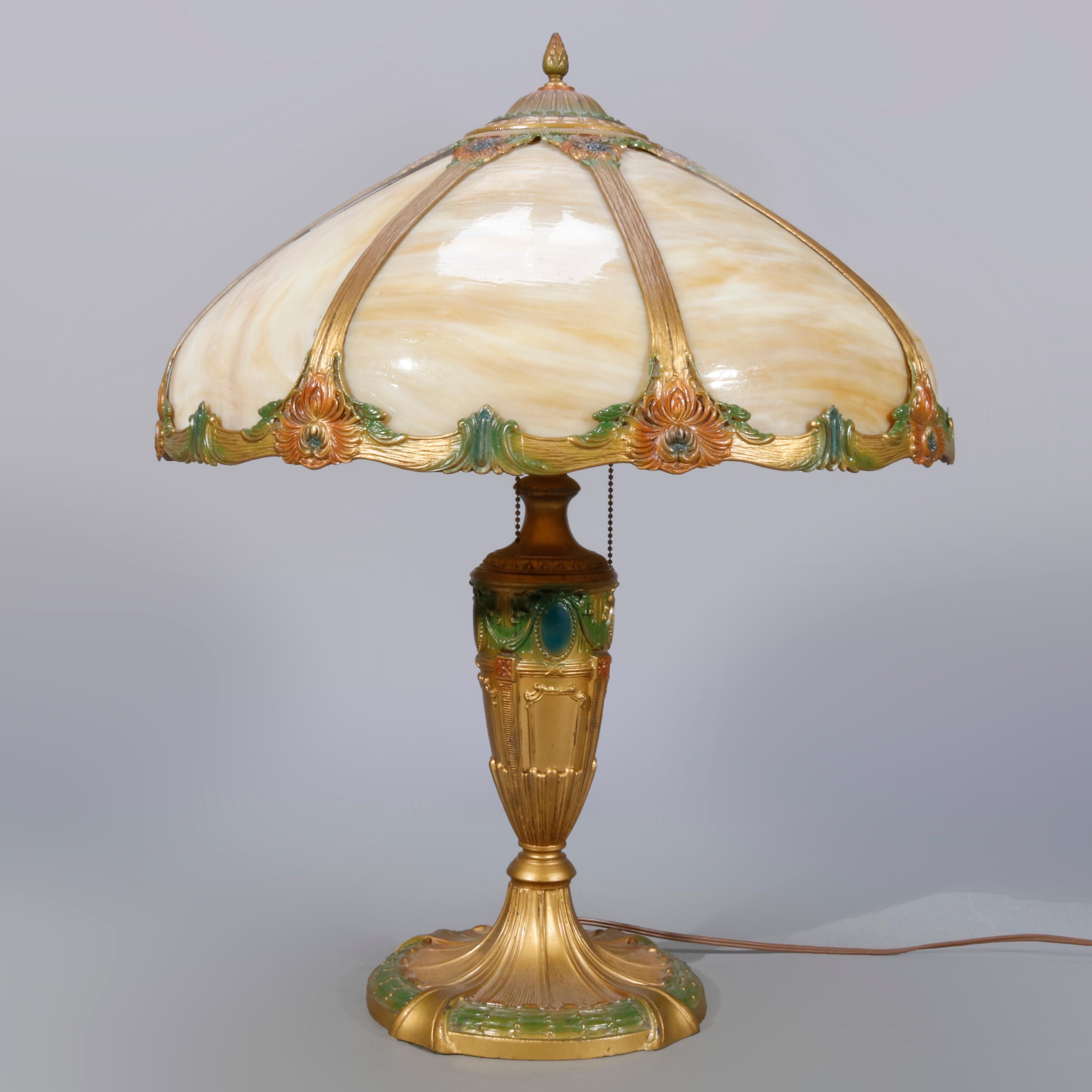An antique Arts & Crafts table lamp offers domed shade with curved slag glass panels over double socket cast urn form base with polychromed floral elements, 