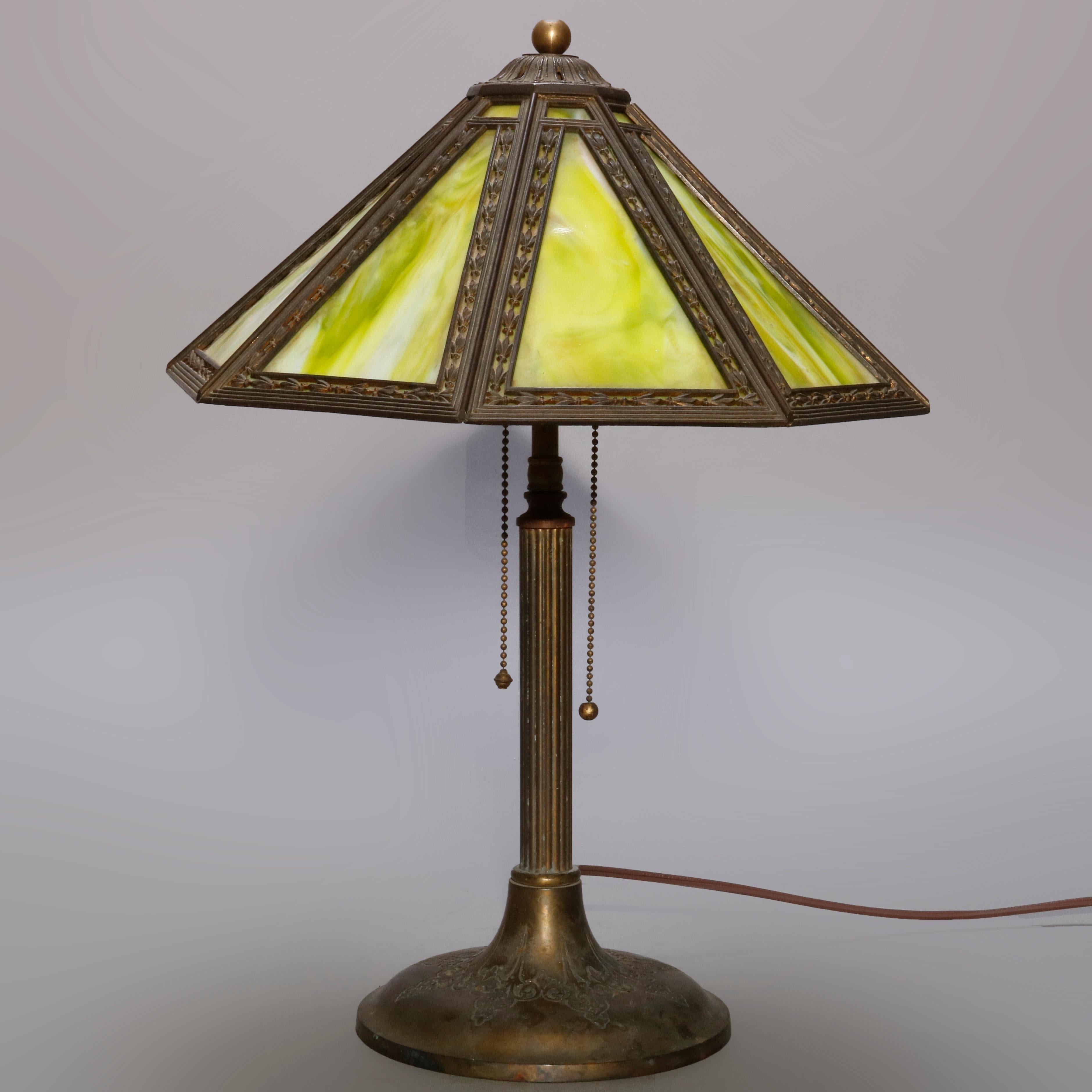 An antique Arts & Crafts table lamp by Bradley and Hubbard offers paneled shade having pierce bellflower frame and housing green slag glass over double socket base with reeded column and seated on base with foliate scroll decoration, circa