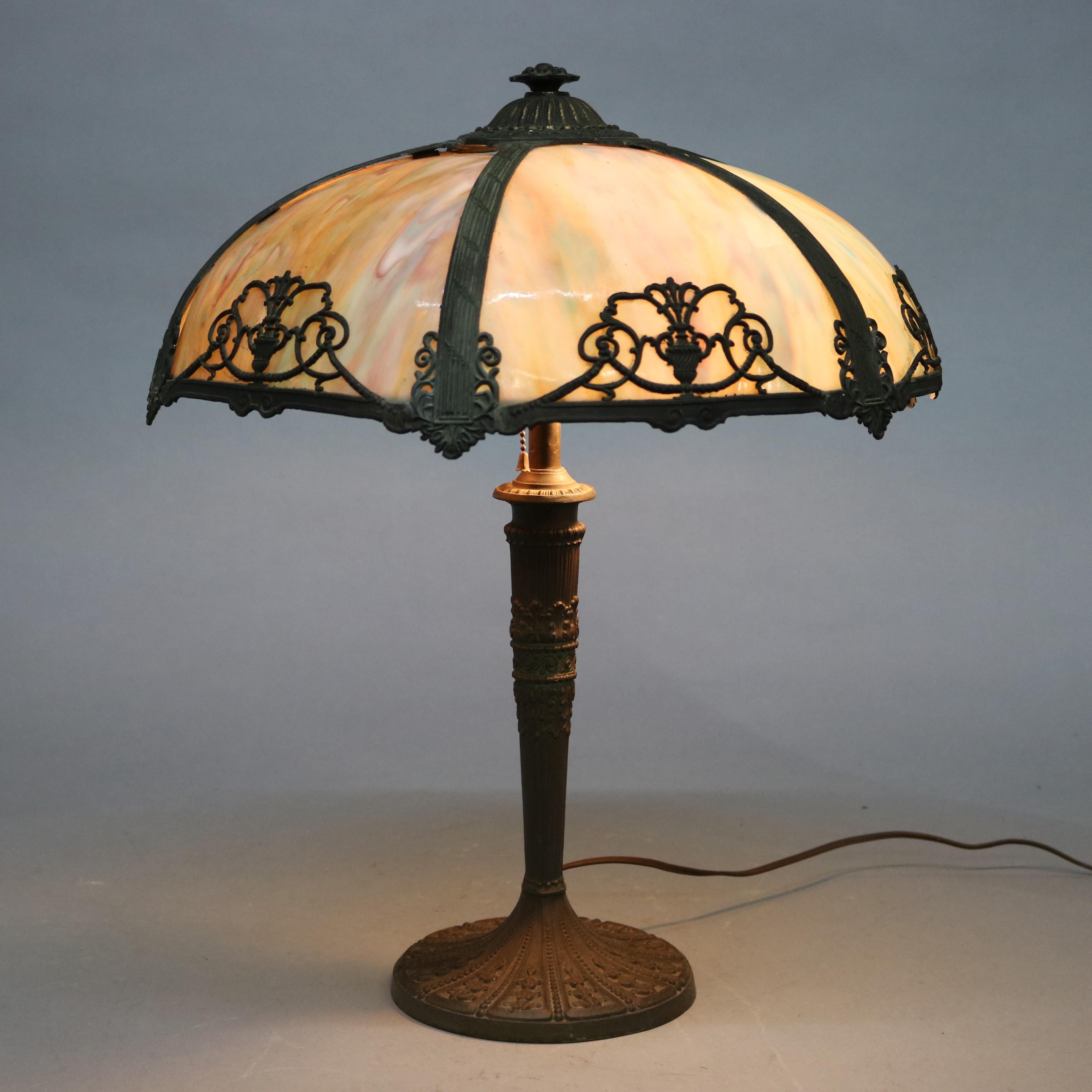 An Arts & Crafts table lamp in the manner of Bradley and Hubbard offers cast frame with urn and foliate elements and housing bent slag glass surmounting double socket base, circa 1920.

Measures: 22