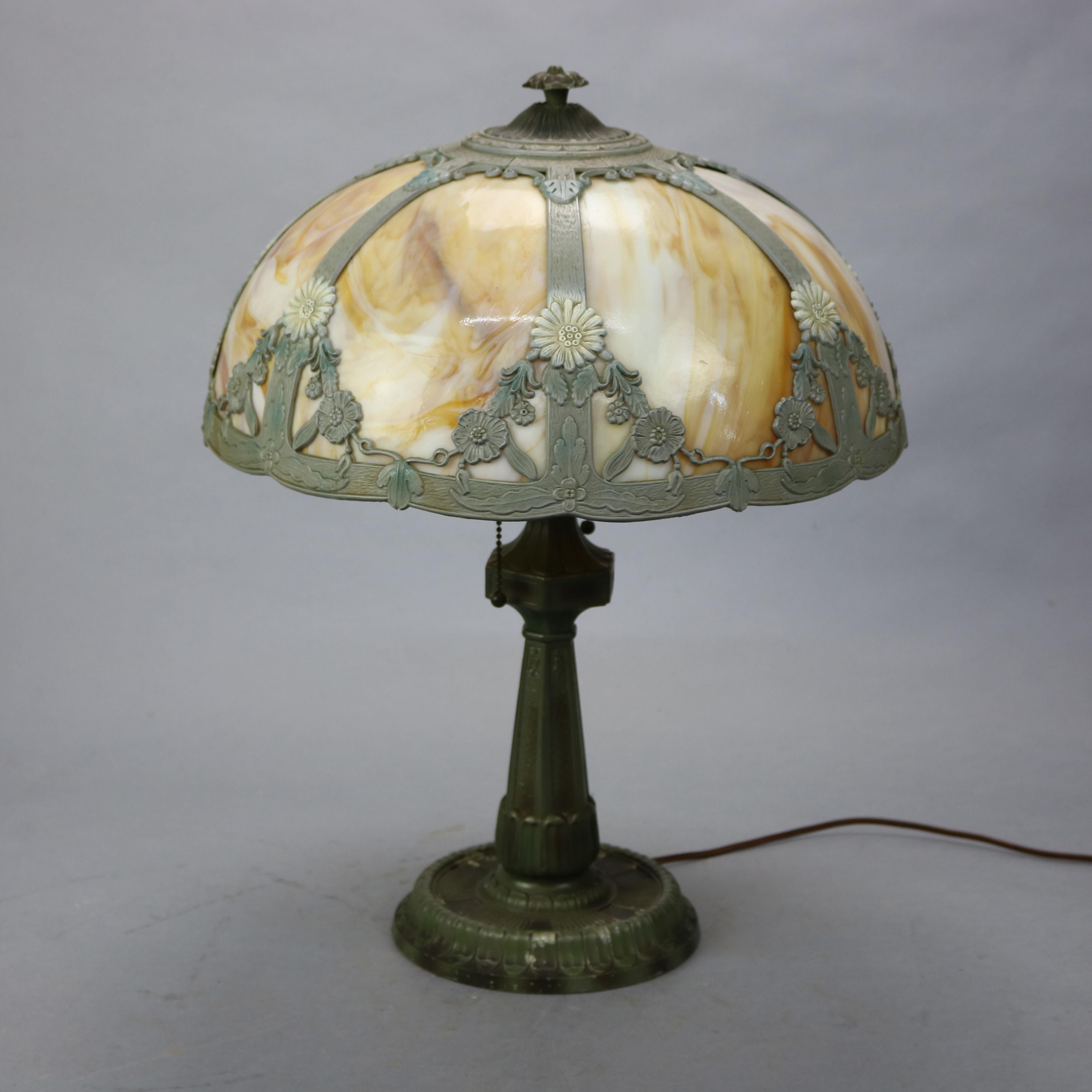 An antique Arts and Crafts table lamp in the manner of Bradley and Hubbard offers dome form shade with cast shade having stylized urn and foliate elements and housing bent slag glass panels over double socket cast metal base, c1920

Measures -