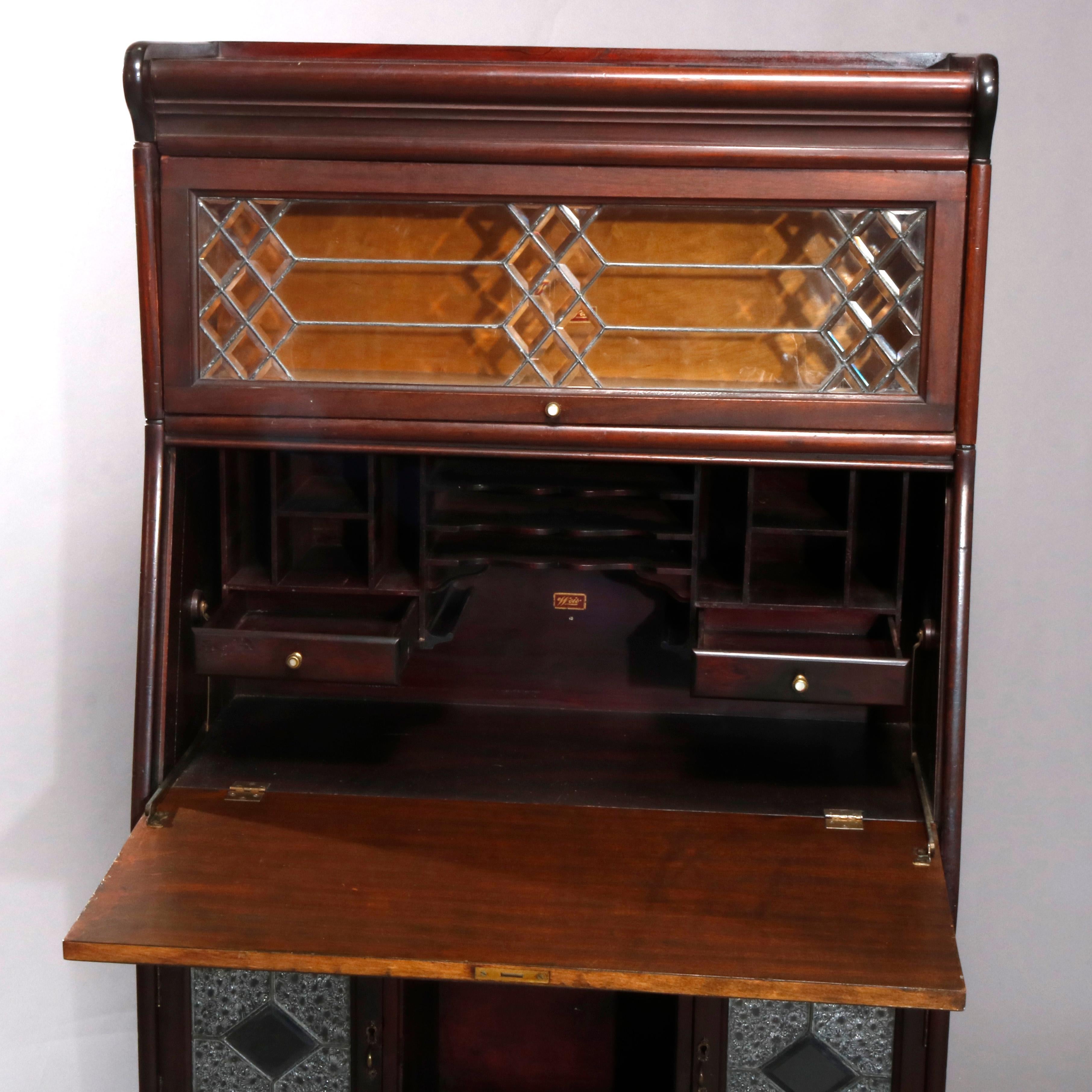 An antique Arts & Crafts secretary by Weis offers mahogany construction with upper leaded beveled glass Barrister bookcase surmounting slant front secretary over case having frieze and lower convex drawers sandwiching pigeon hole with flanking