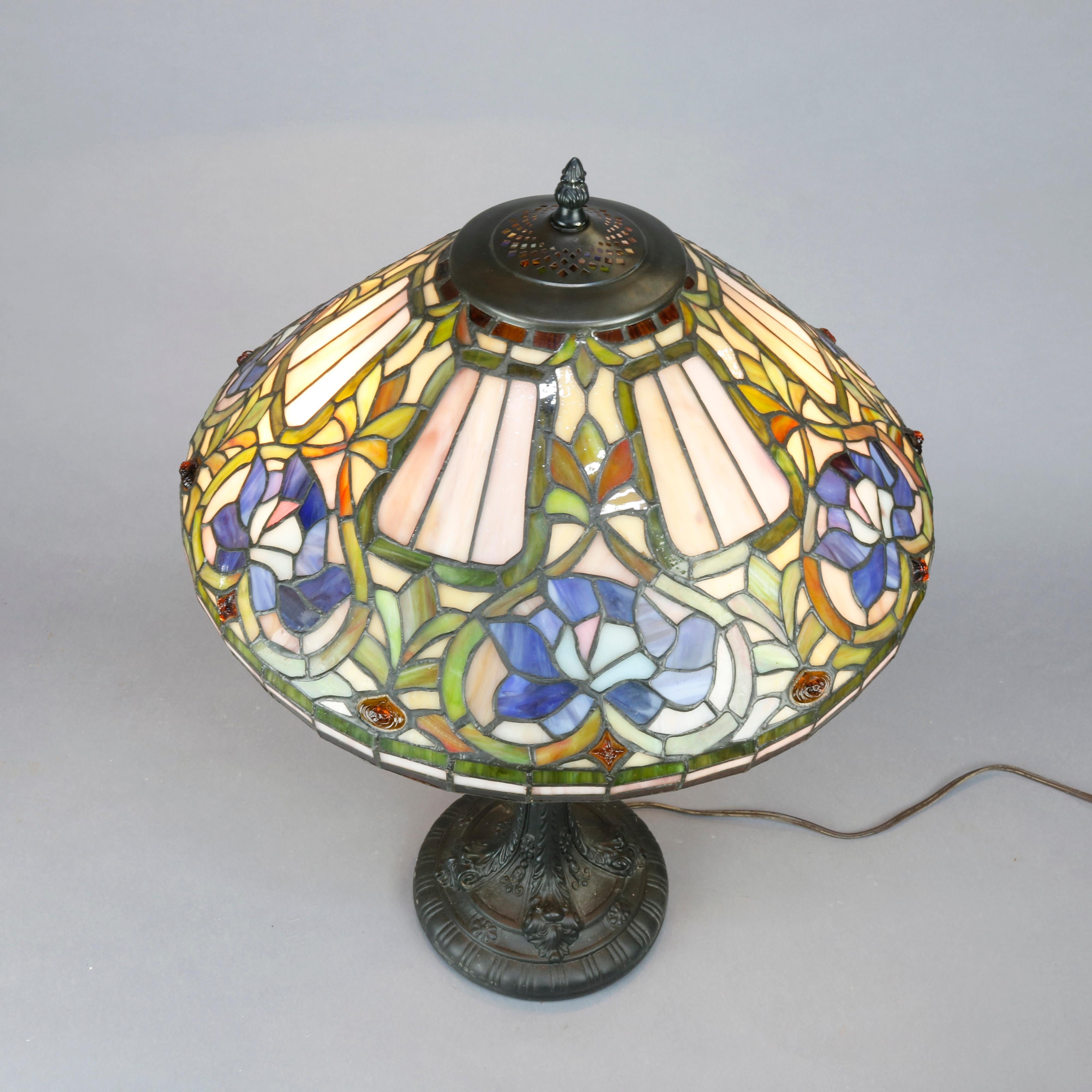 An antique Arts & Crafts table lamp in the manner of Tiffany Studios offers leaded slag and stained glass shade having stylized floral design with molded glass jewels surmounting foliate double cast double socket base, 20th century.

Measures: 26