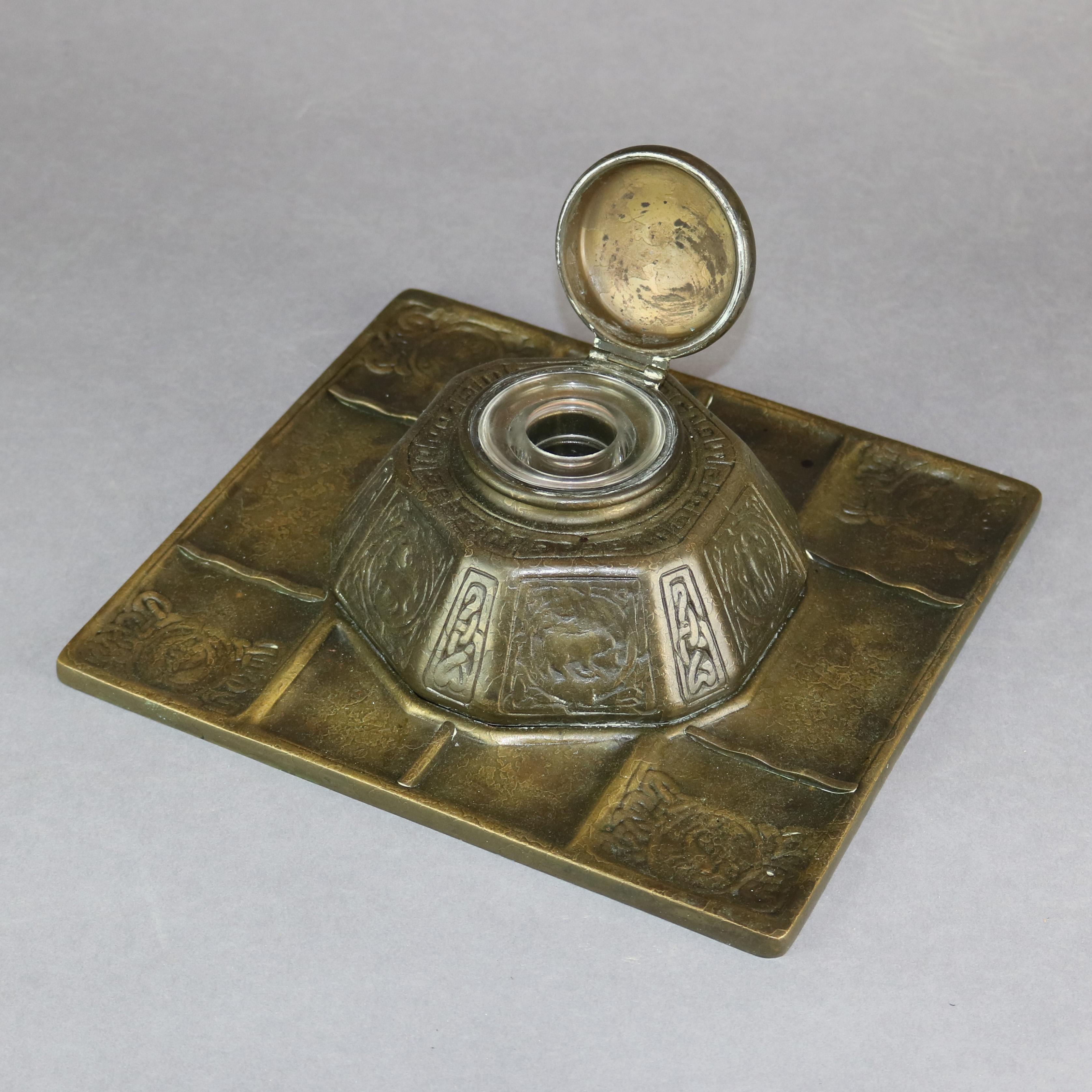 An antique inkwell by Tiffany Studios offers cast bronze form in pyramidal form having alternating panels of Zodiac signs and stylized interlocking chains and seated on segmented tray base, lid opens to glass ink vessel, signed 