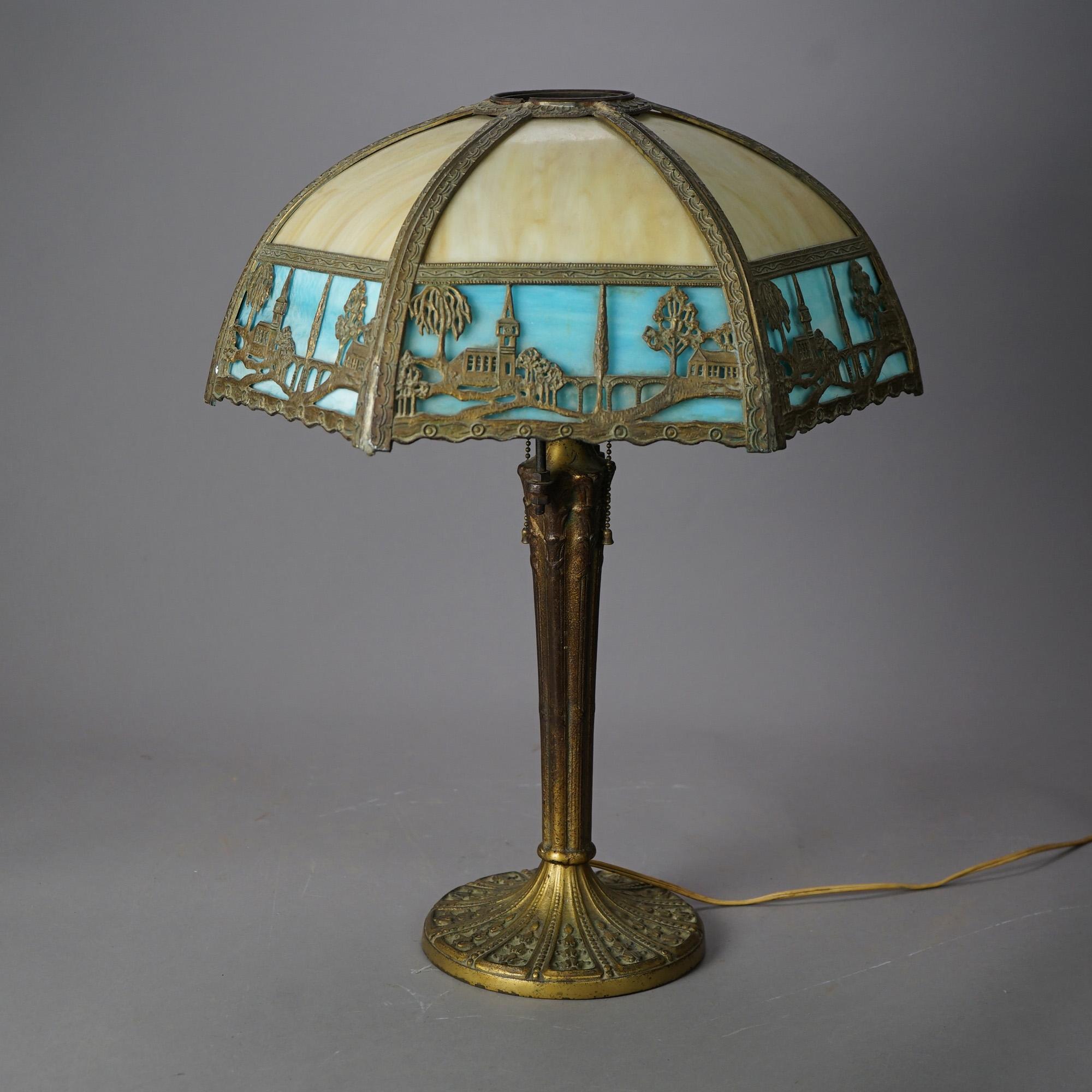 An antique Arts and Crafts table lamp offers dome form shade having cast metal frame with band having countryside scene with a church and housing two-tone bent slag glass panels over double socket cast base, c1920

Measures - 22