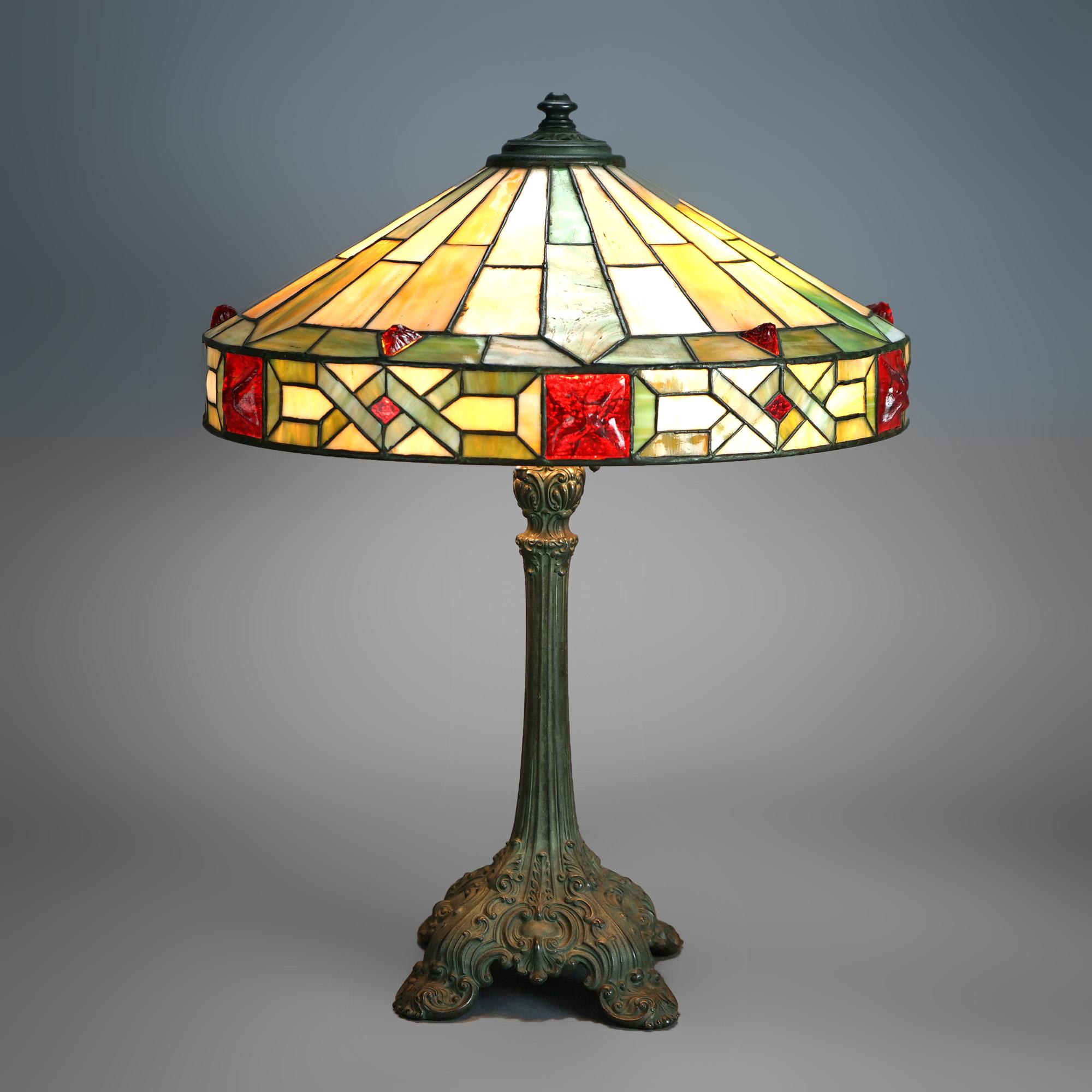 An antique Arts & Crafts table lamp by Wilkinson offers leaded slag, chunk and jewel glass shade in geometric form over triple socket cast base having scroll and foliate elements raised on stylized feet, c1920

Measures- 24''H x 18''W x 18''D