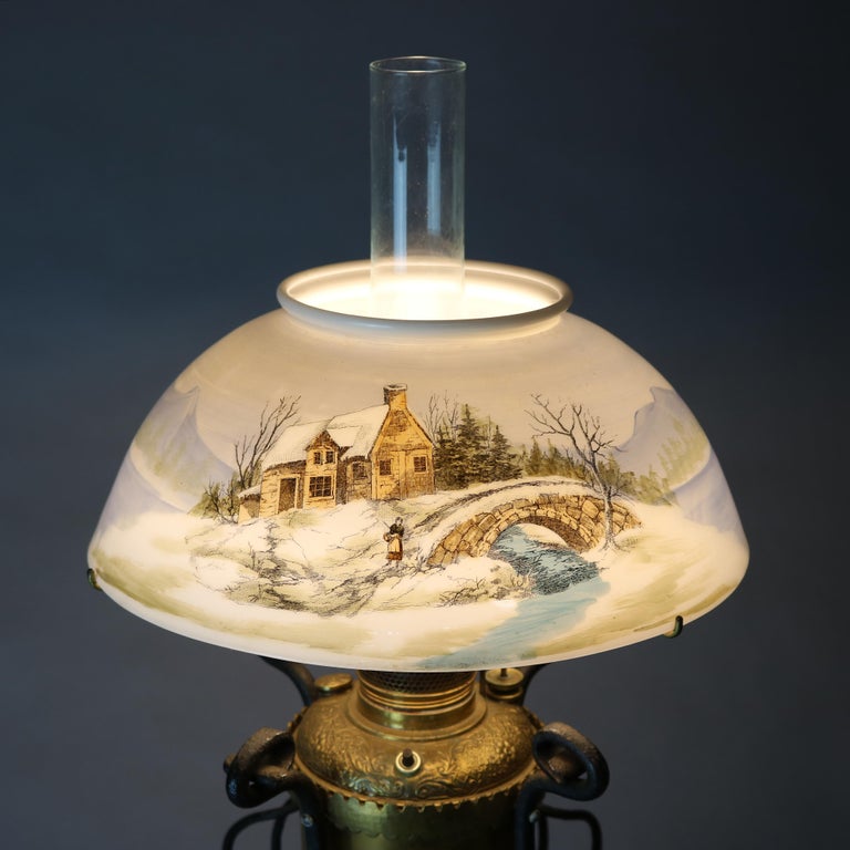 An antique Arts & Crafts table lamp offers brass urn form font having foliate and scroll embossed upper, seated in wrought iron frame with scroll feet and surmounted by glass shade with hand painted winter landscape scene having structure and