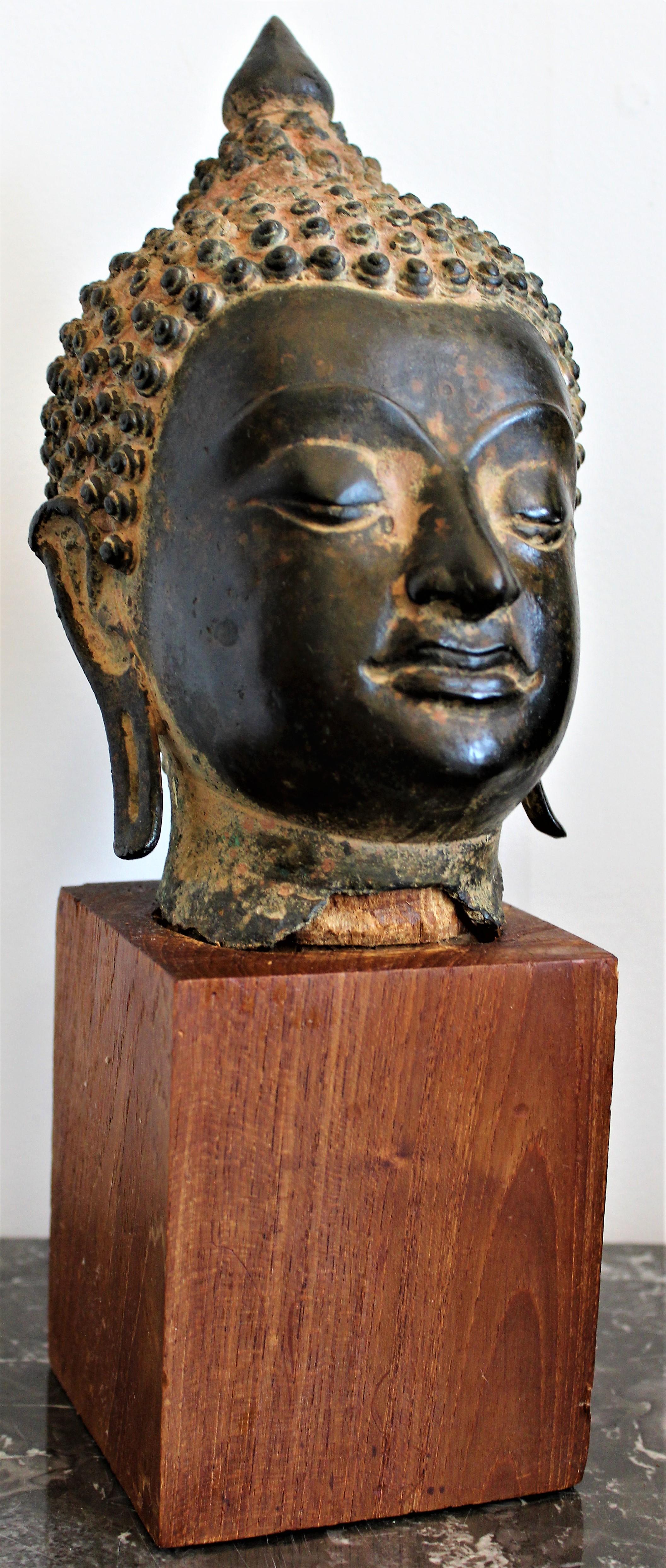 Antikes asiatisches und äußerst detailreiches Bronzegussfragment eines Buddhakopfes aus dem 18. Jahrhundert, das vermutlich thailändischen oder tibetischen Ursprungs ist. Das Skulpturenfragment ist sicher auf einem einfachen Teakholzblock