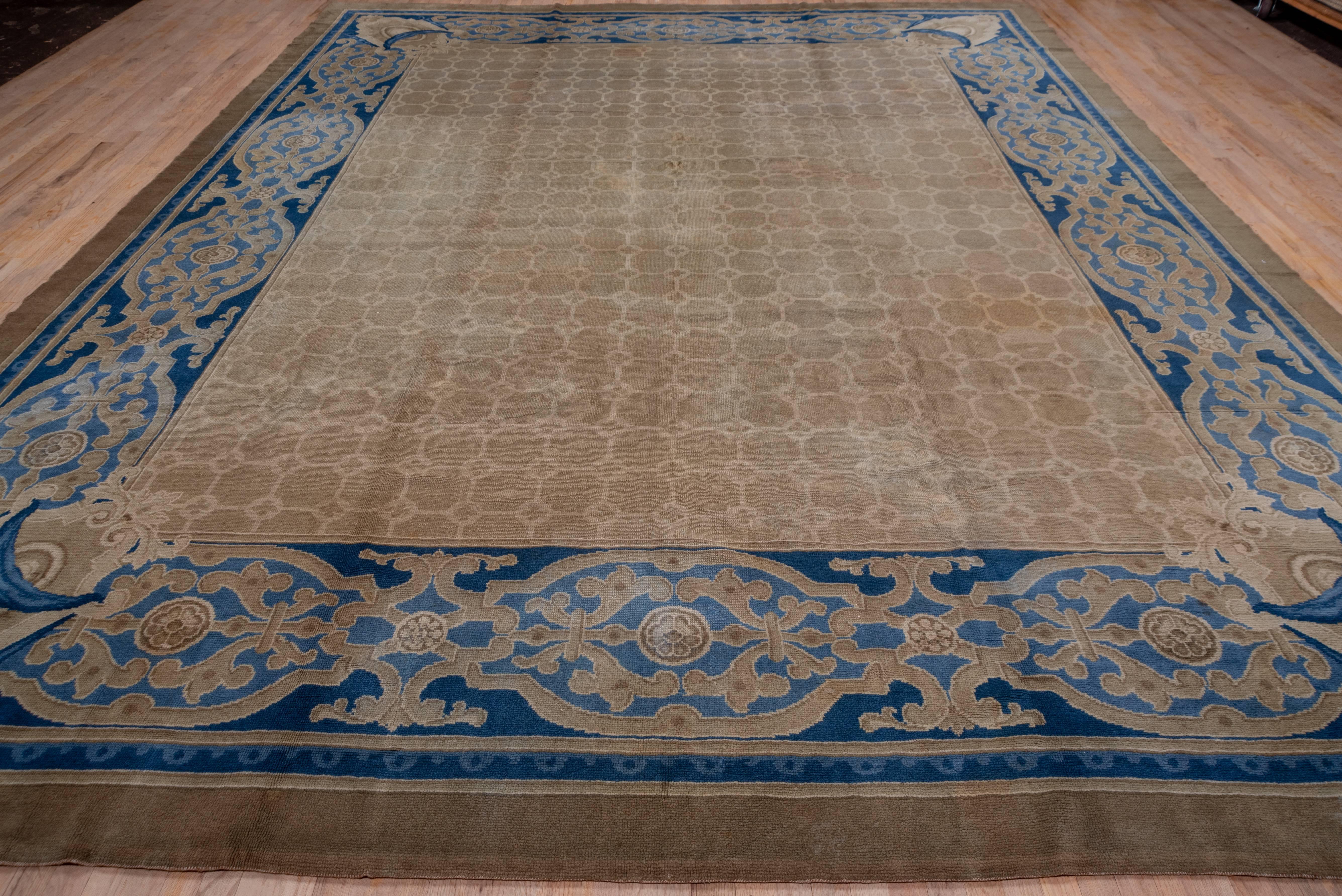 This thick pile carpet has a tan field with a small all-over repeating pattern of fat eight pointed brown tiles on the beige-tan field. A blue-slate border of joined cartouches with contoured strapwork makes an appropriate frame. The drapery swag