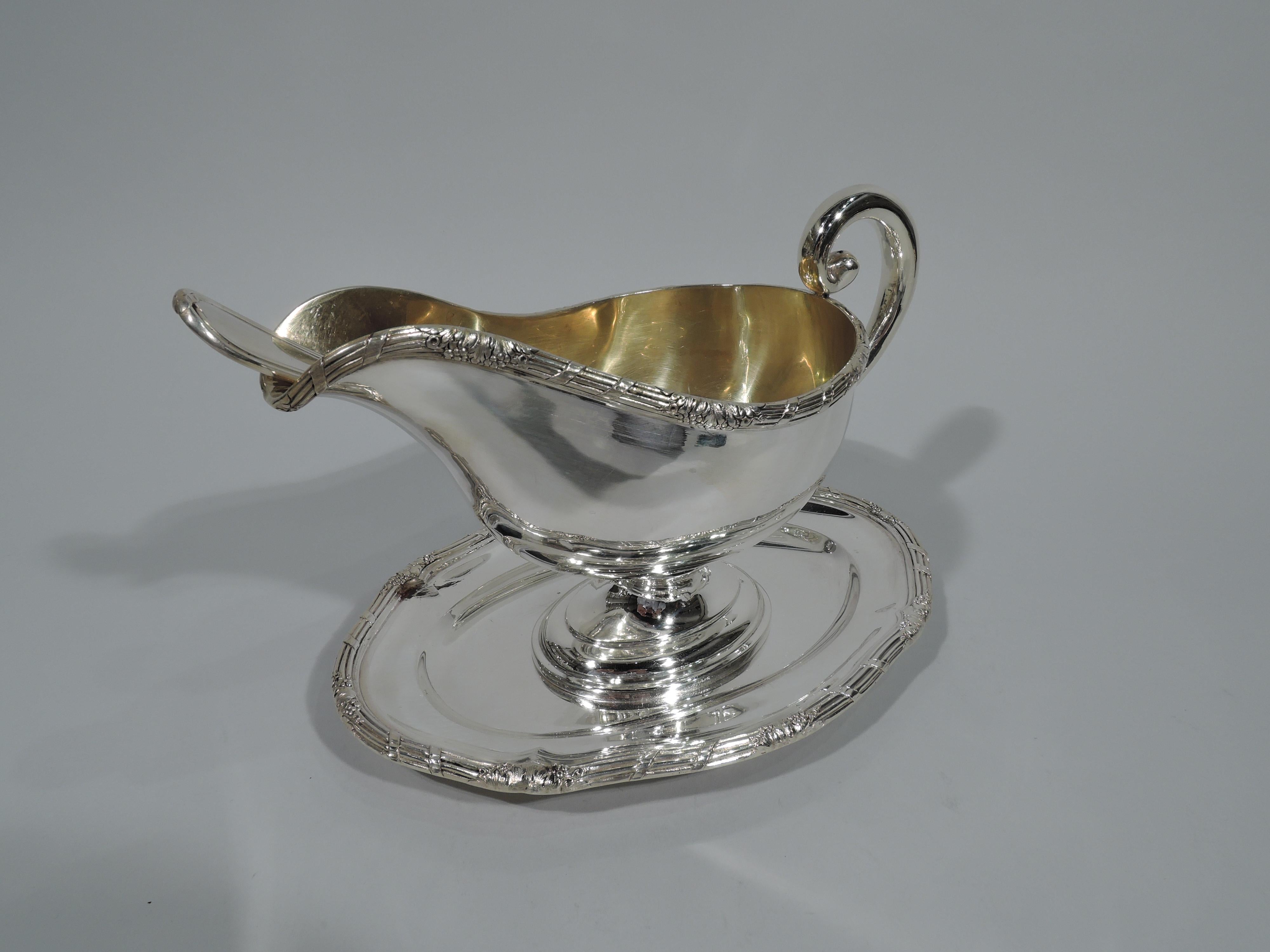 Austrian neoclassical 800 silver gravy boat on stand with ladle, circa 1920. Boat: Helmet mouth with high-looping scroll handle, stepped oval foot, gilt-washed interior, and reeded rim with leaves and berries. Stand: Shaped oval with same rim. Ladle