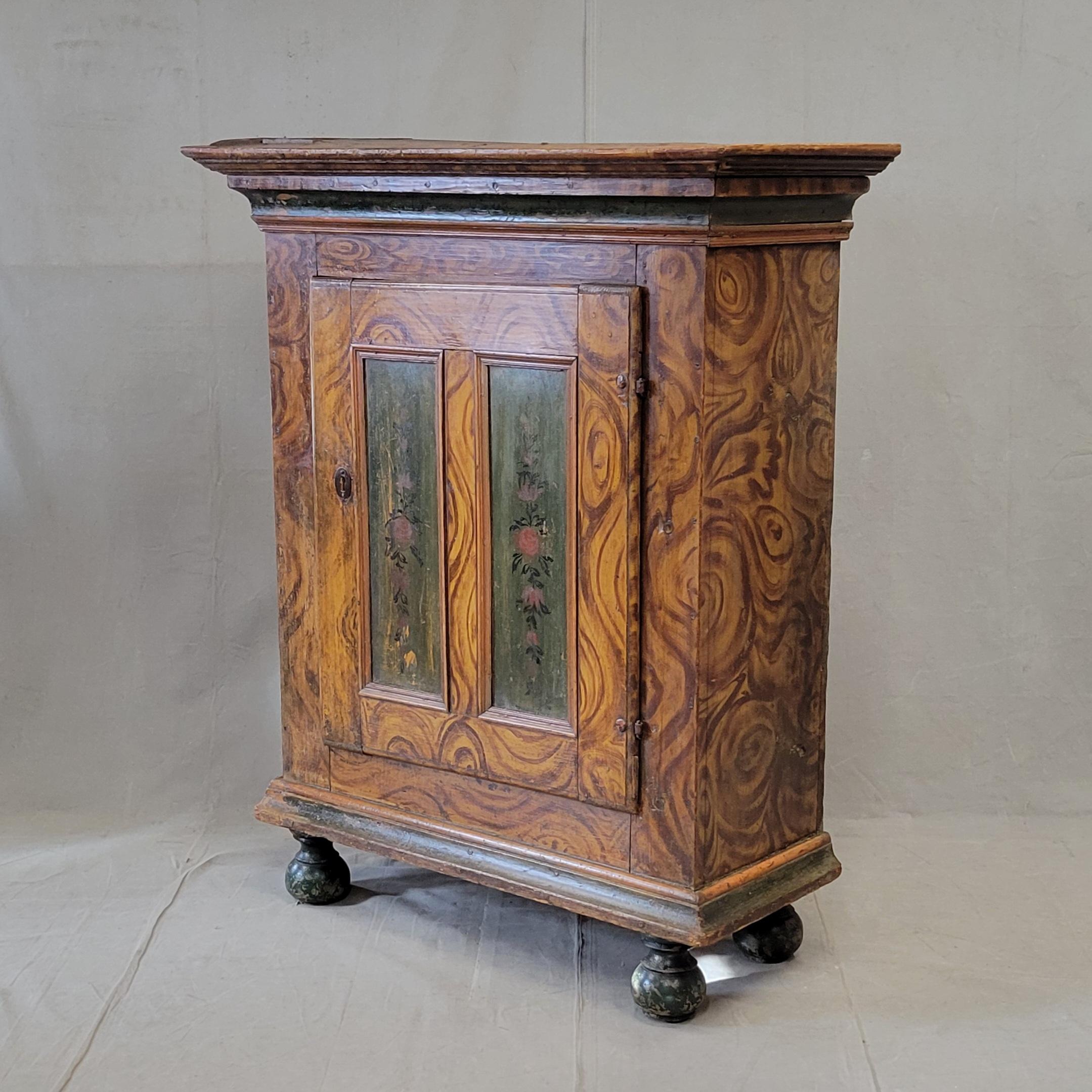 Encantador y funcional armario antiguo austriaco o alemán de finales del siglo XIX, pintado a la veta (faux bois) con toques rojos y azules/verdes en un motivo floral en los paneles de la puerta delantera. El interior tiene un estante para guardar
