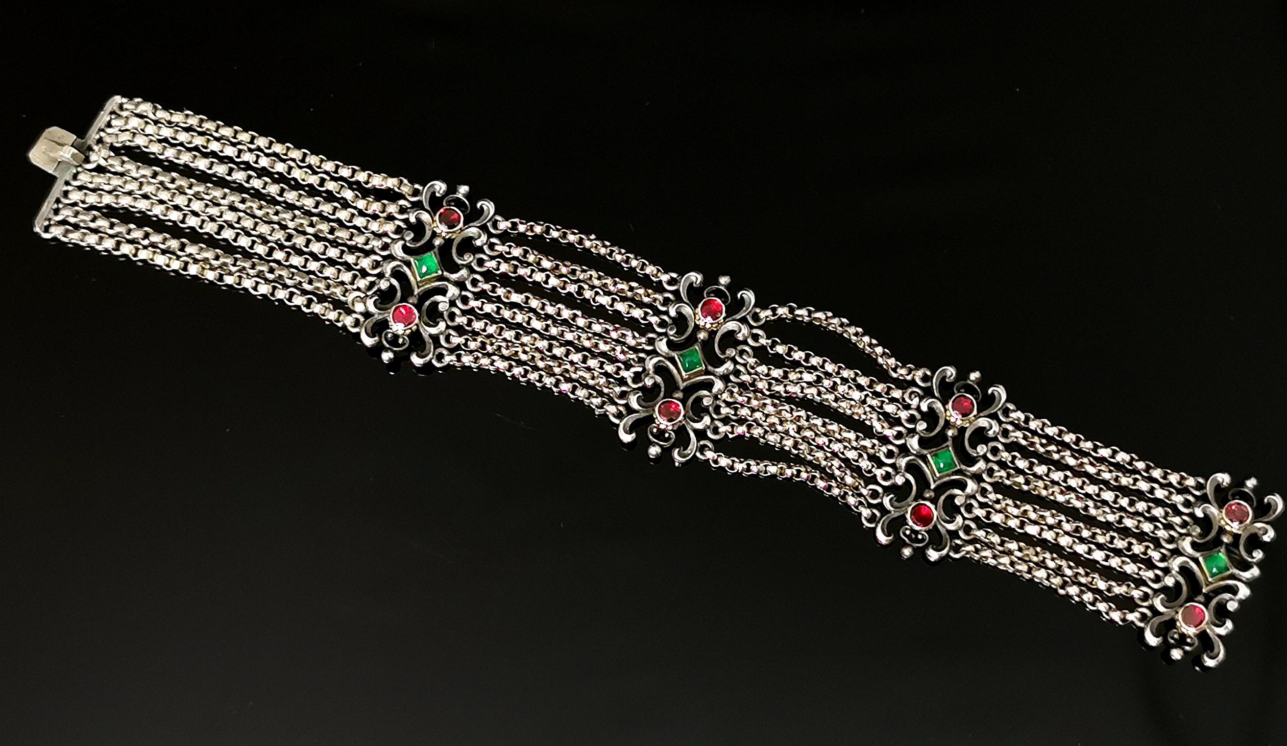 A gorgeous antique Austro-Hungarian bracelet.

This is a multi-strand bracelet with eight rows of silver rolo or belcher chain joined together with jewelled, scrolling silver panel spacers.

The bracelet is set with rich red garnets and green