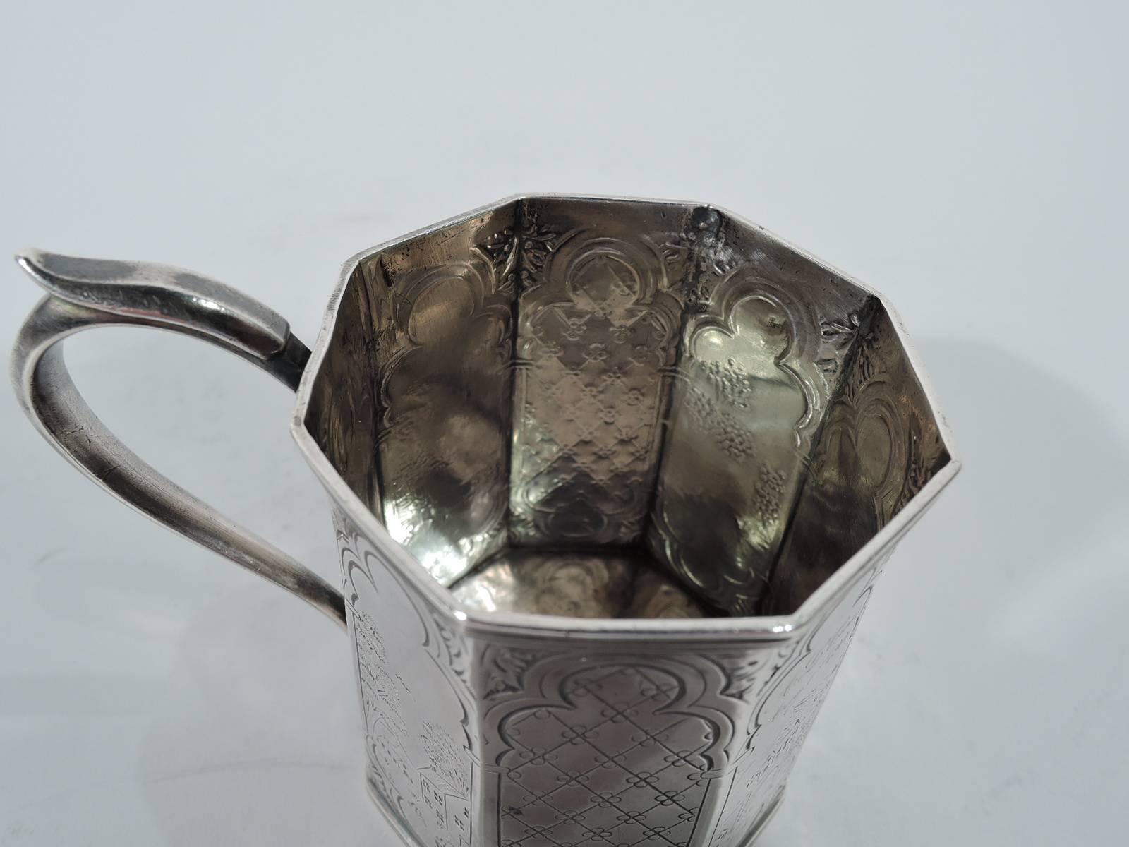 Pastoral coin silver christening mug. Faceted and upward tapering sides with capped scroll handle. Engraved ornament: Tubular multifoils of which some inset with landscapes and country cottages and others with diaper ornament. Two have engraved