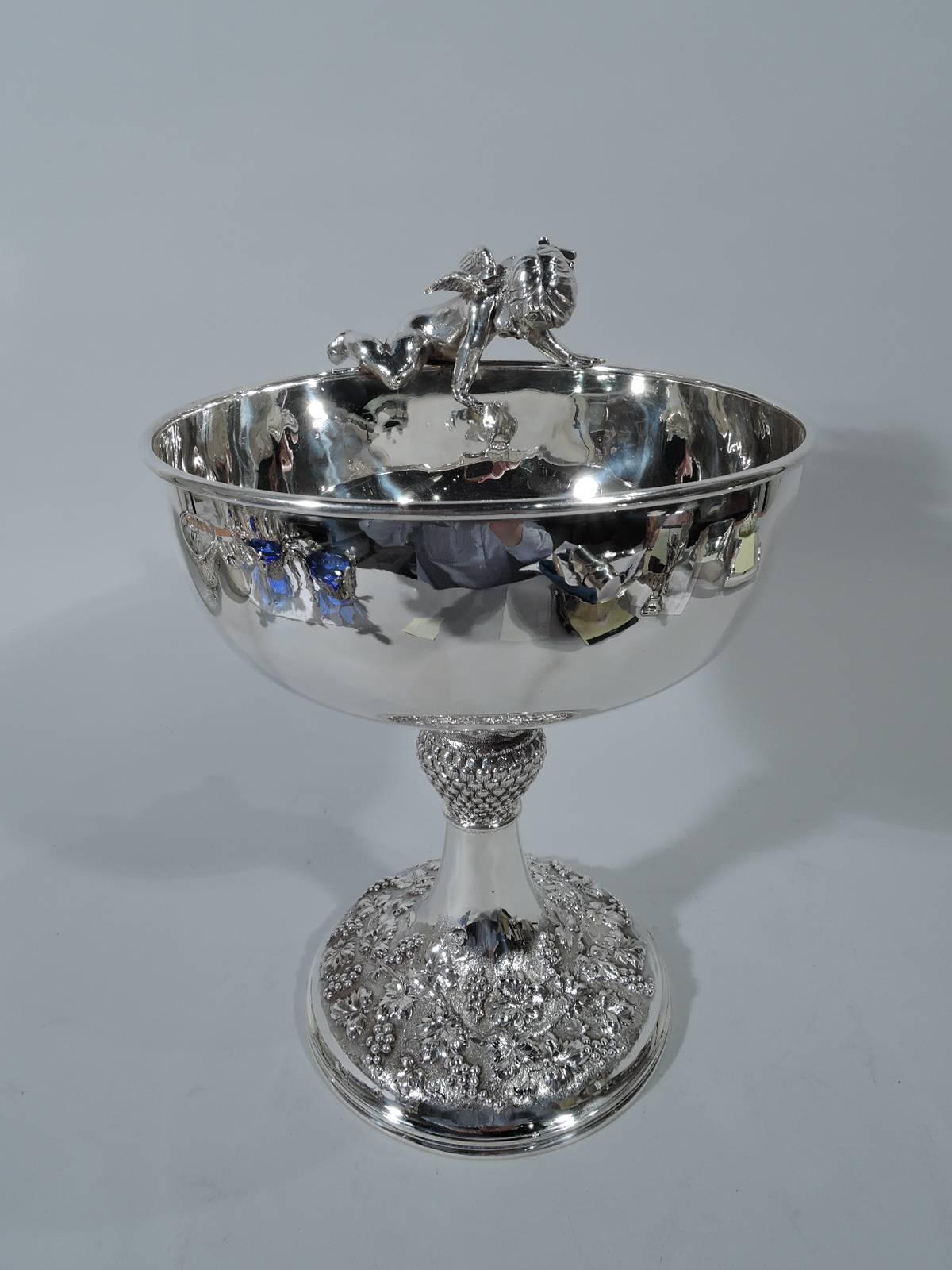 Edwardian sterling silver golf trophy. Round bowl on conical support with pineapple knop. Raised round foot decorated with chased fruiting grapevine. Precariously perched on rim is cast cupid figure. A chubby thigh hangs over the rim while a hand