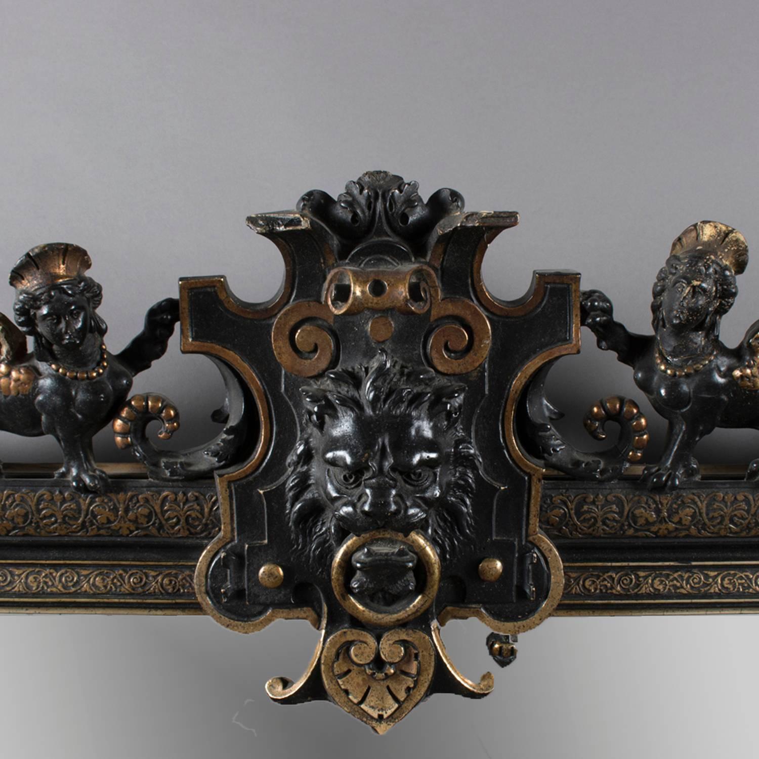 Large antique Baroque figural over mantel mirror features ebonized and carved scroll and foliate decorated frame with pierced crest having central lion mask with flanking Greek Sphinx and pierced scroll form apron, gilt accents throughout and