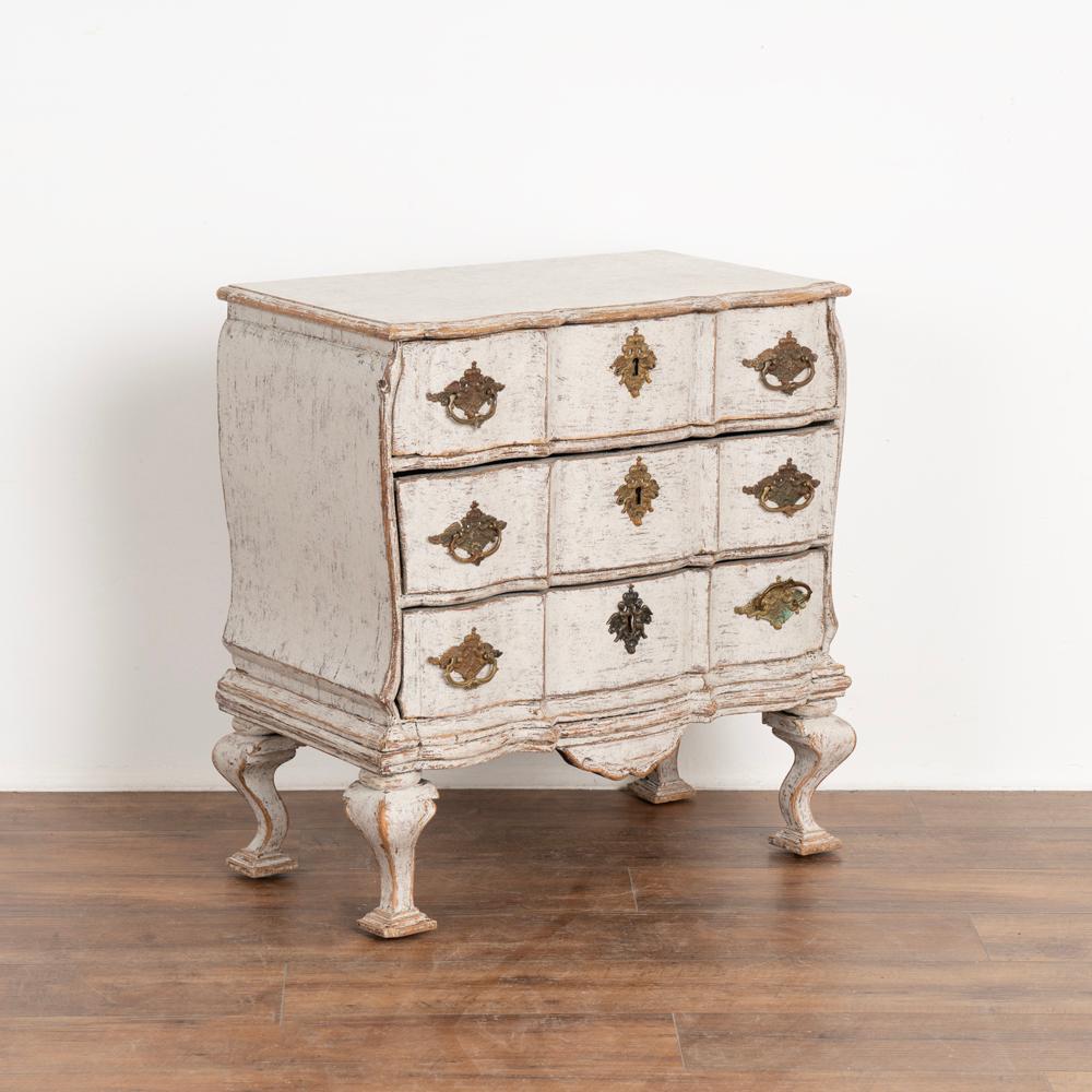 A decorative small Swedish Baroque oak chest of drawers or nightstand resting on cabriolet feet.
Painted in a professionally applied newer white layered finish, softly distressed.
Three functioning drawers each with two brass pulls. One key