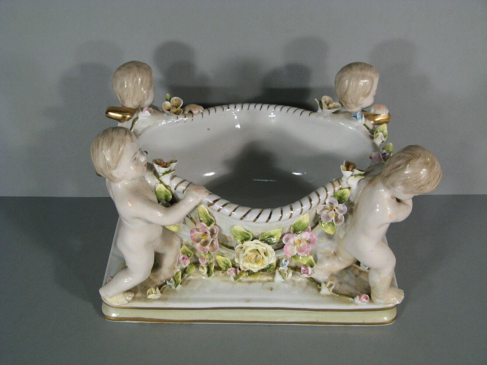 This highly decorative antique centerpiece, crafted in polychrome porcelain, showcases a stunning Baroque style. The rectangular base supports a bowl carried by four cherubic putti, adorned with garlands of flowers. A mark, imitating the renowned